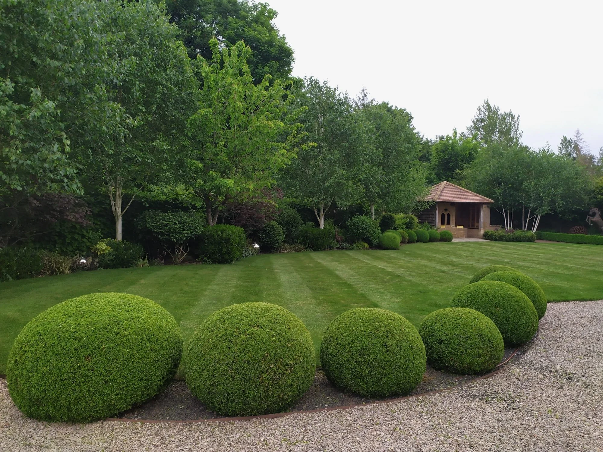 A well-manicured garden with large, round, green bushes in the foreground, a lush green lawn, several trees, and a small pavilion with a tiled roof in the background.