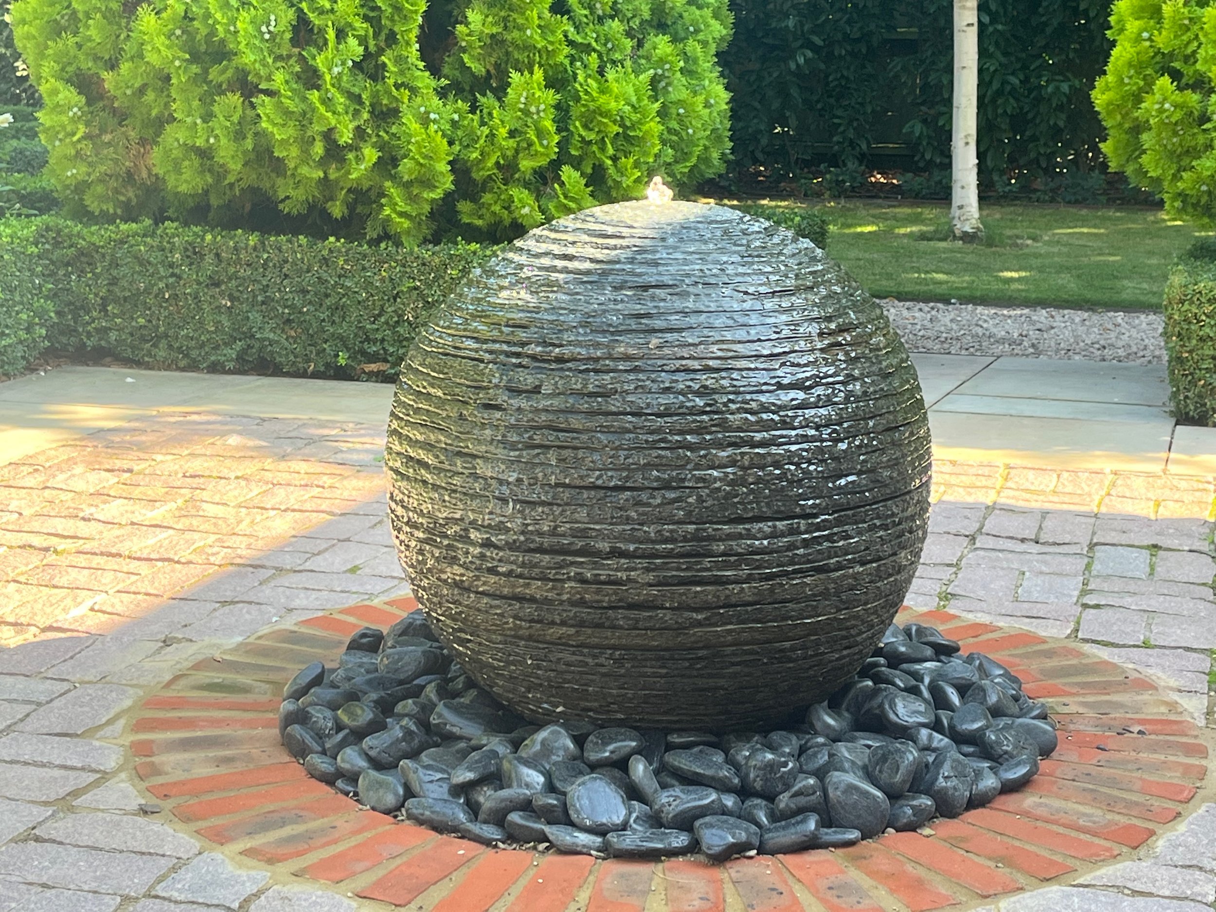 A rounded, layered stone fountain with water flowing over its surface, surrounded by smooth black stones, situated on a brick and concrete patio in a garden with greenery and trees.