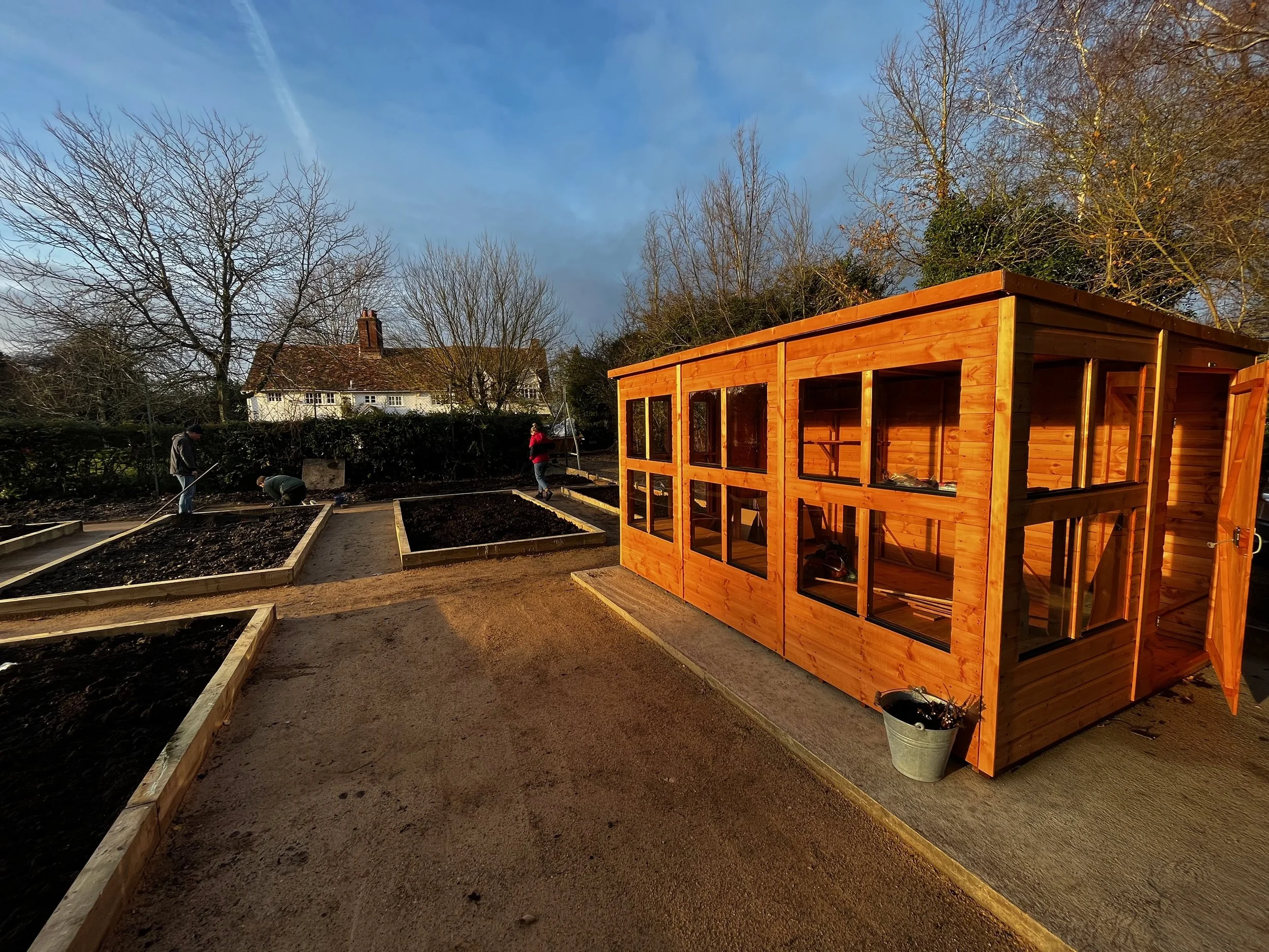 A group of people planting a garden near a wooden house, with trees and a blue sky in the background.