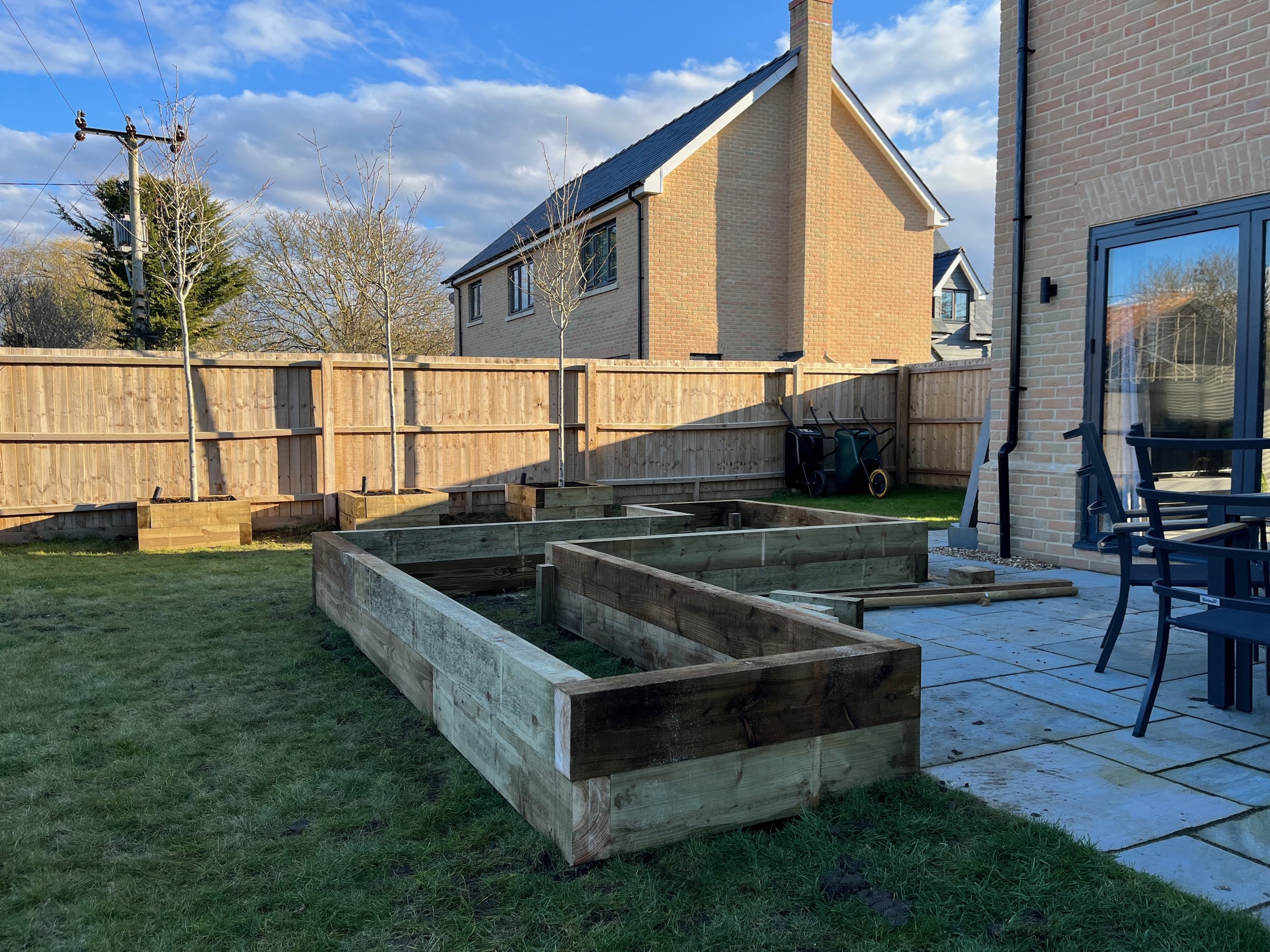 A backyard with a wooden fence, raised garden beds, a lawn, and a house with brick walls. There are two leafless trees, gardening tools, and a wheelbarrow. The sky is partly cloudy with blue sky visible.