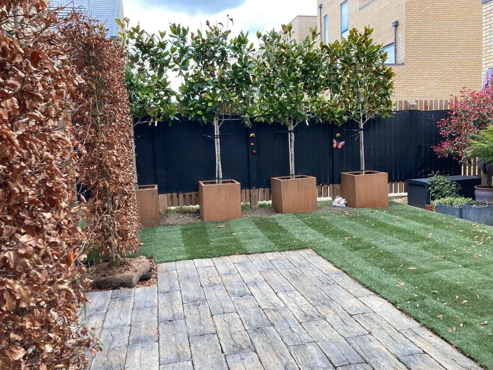 Backyard garden with potted trees, a brick patio, artificial grass, and a black fence with decorations, surrounded by various plants and a brown fence in the background.