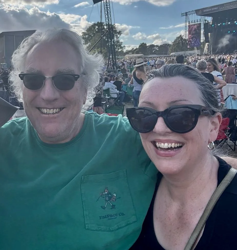 Two smiling people wearing sunglasses at an outdoor music festival during the day, with a stage, crowd, and cloudy sky in the background.