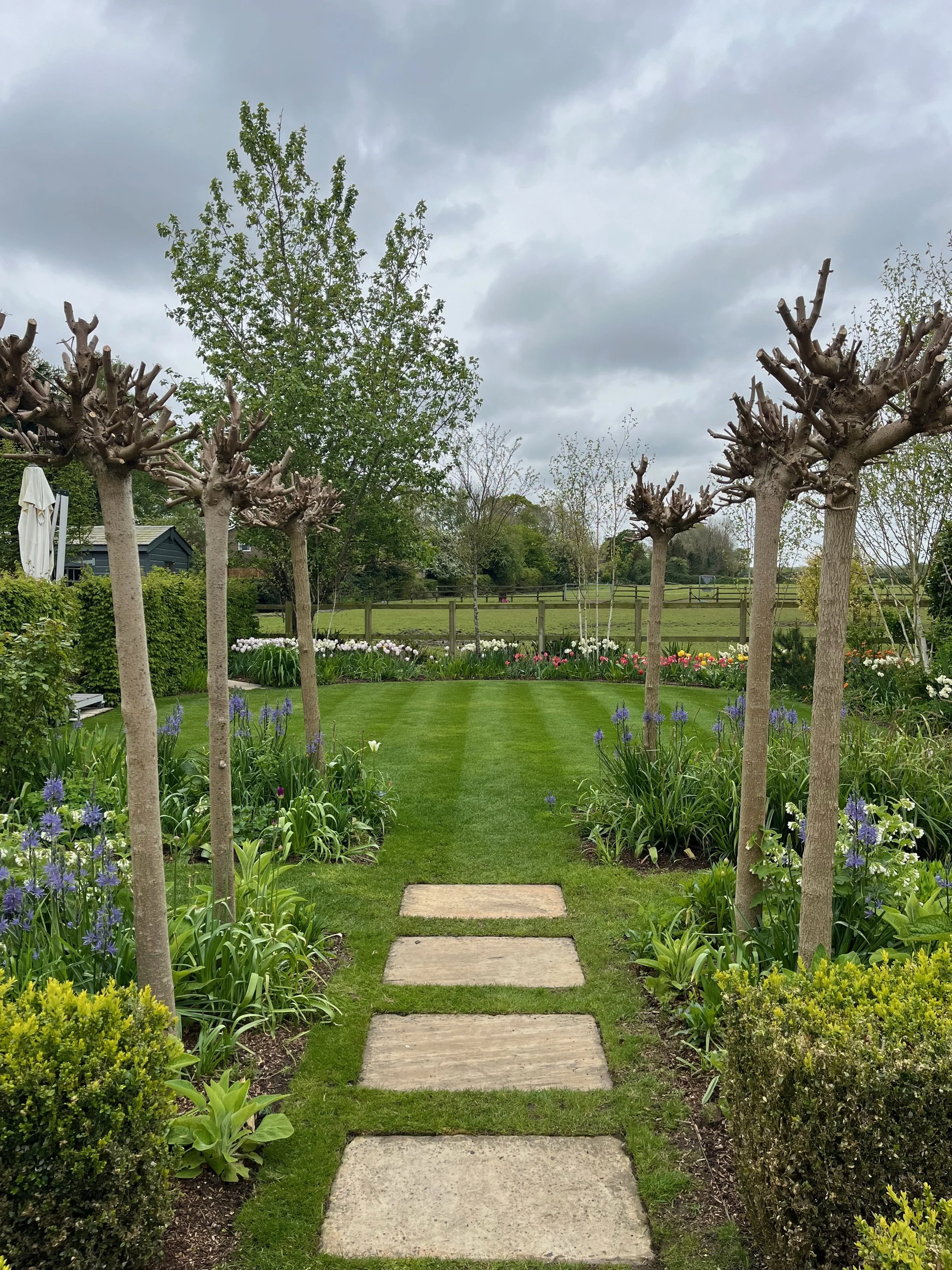 A garden pathway with rectangular stone stepping stones leading through flower beds and trimmed bushes, with pruned trees on either side, green lawn, and cloudy sky overhead.