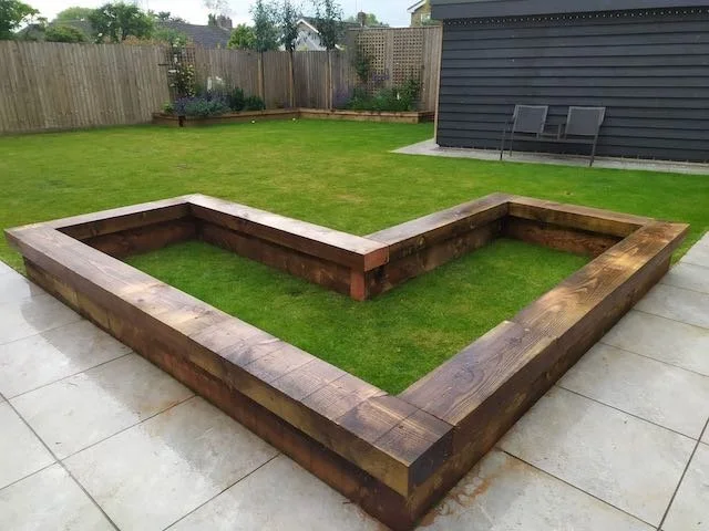 Backyard with a raised garden bed outline made of wooden planks, surrounded by grass and a concrete patio.