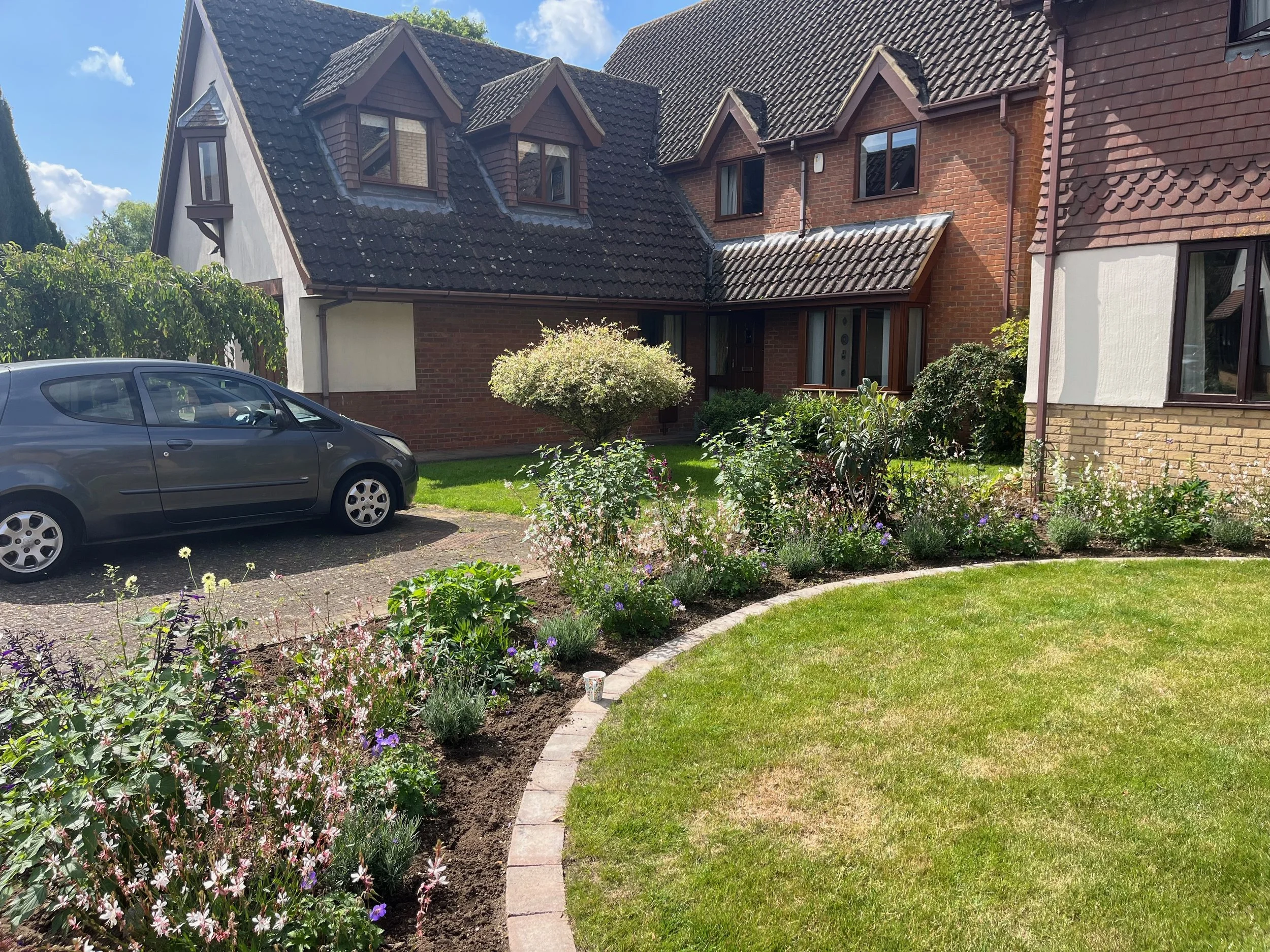 A residential house with a grey tiled roof and brick walls, surrounded by a garden with colorful flowers and shrubs. A silver car is parked on the driveway in front of the house.