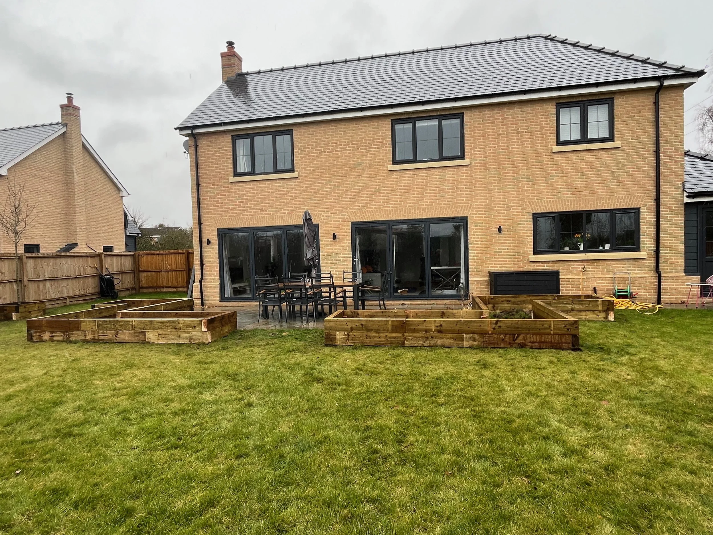 Backyard with a lawn, wooden garden beds, a black patio table with chairs, and a brick house with large windows and a sliding glass door, under an overcast sky.