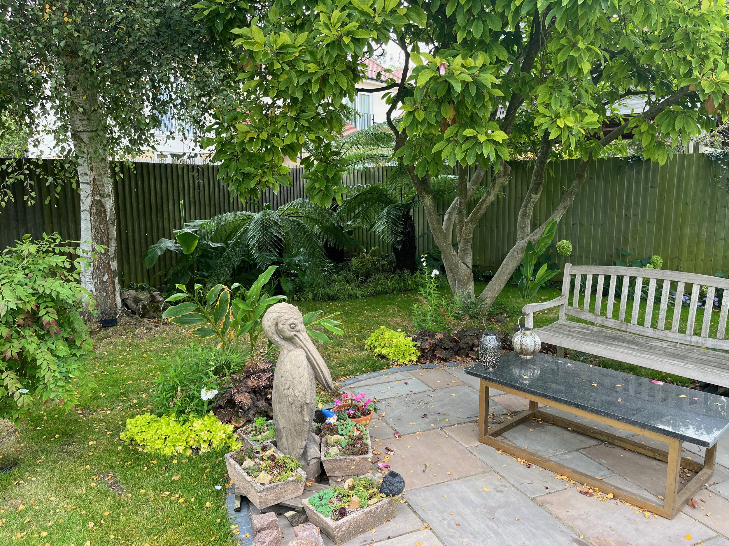 A backyard garden with lush green trees, various plants, and a wooden bench on a paved patio. Decorative lanterns and potted plants are also visible.