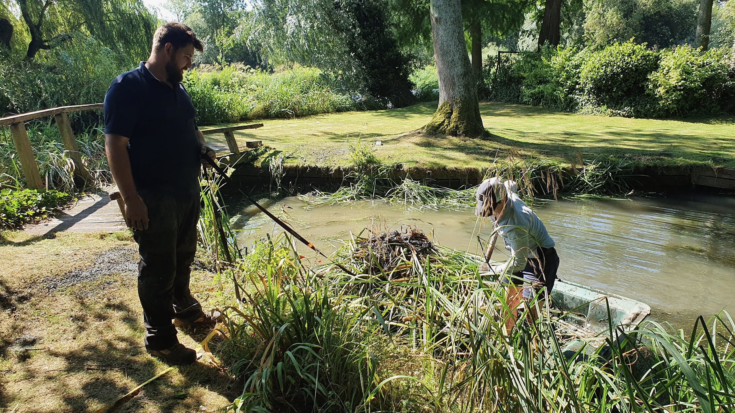 Two men by a pond in a park, with one on the shore and the other in the water, surrounded by trees and greenery.