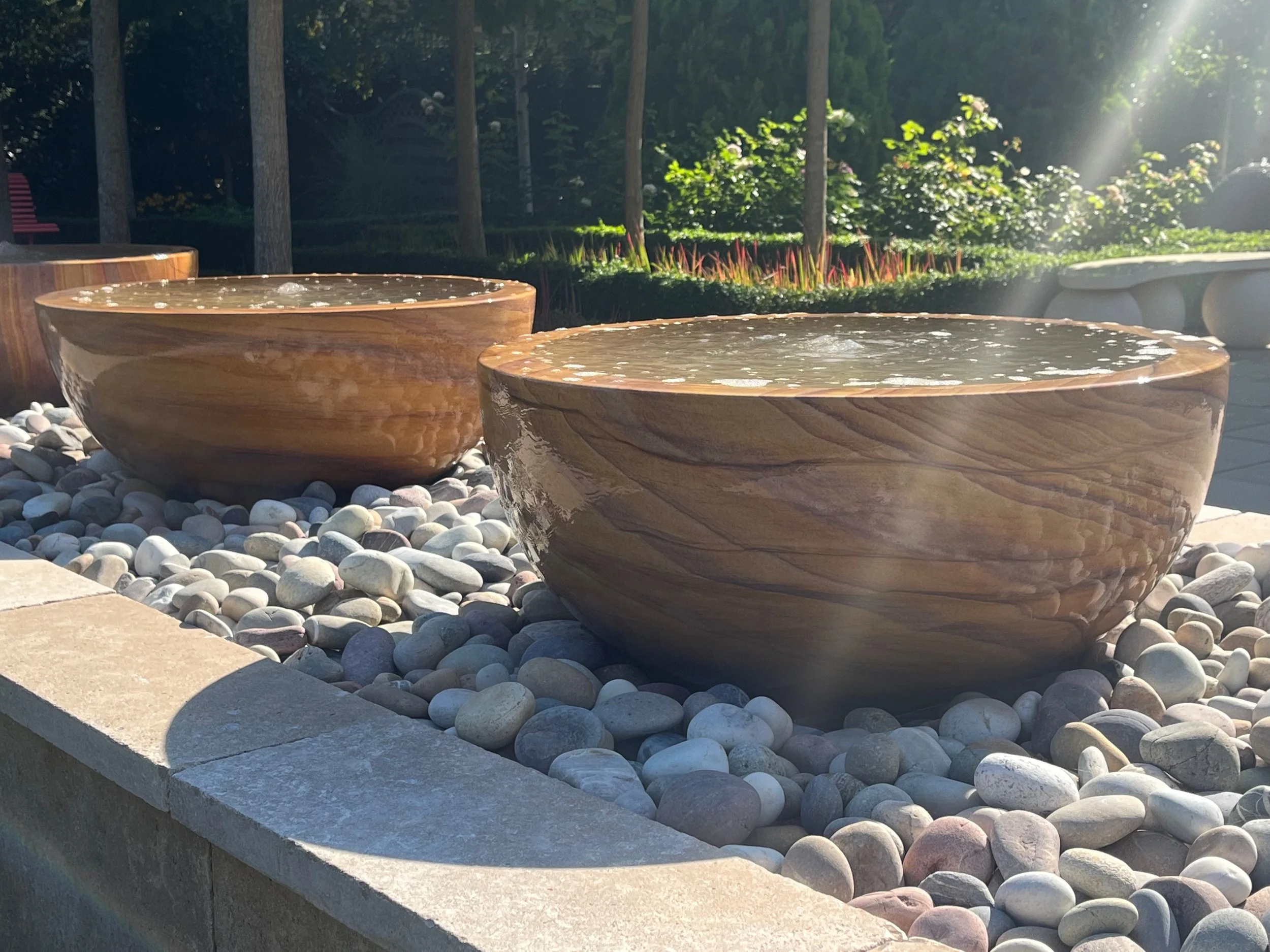 Three wooden bowls filled with water are placed on a stone ledge surrounded by small, smooth white paving stones. In the background, trees, bushes, and a pink park bench are visible in sunlight.