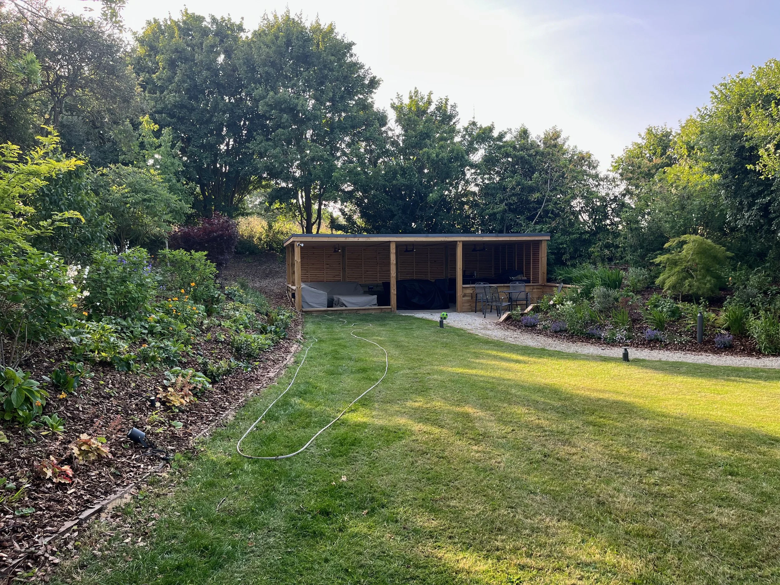 A backyard garden with lush green trees, flowering plants, and a wooden covered patio area with outdoor furniture, a grassy lawn, and garden lights.