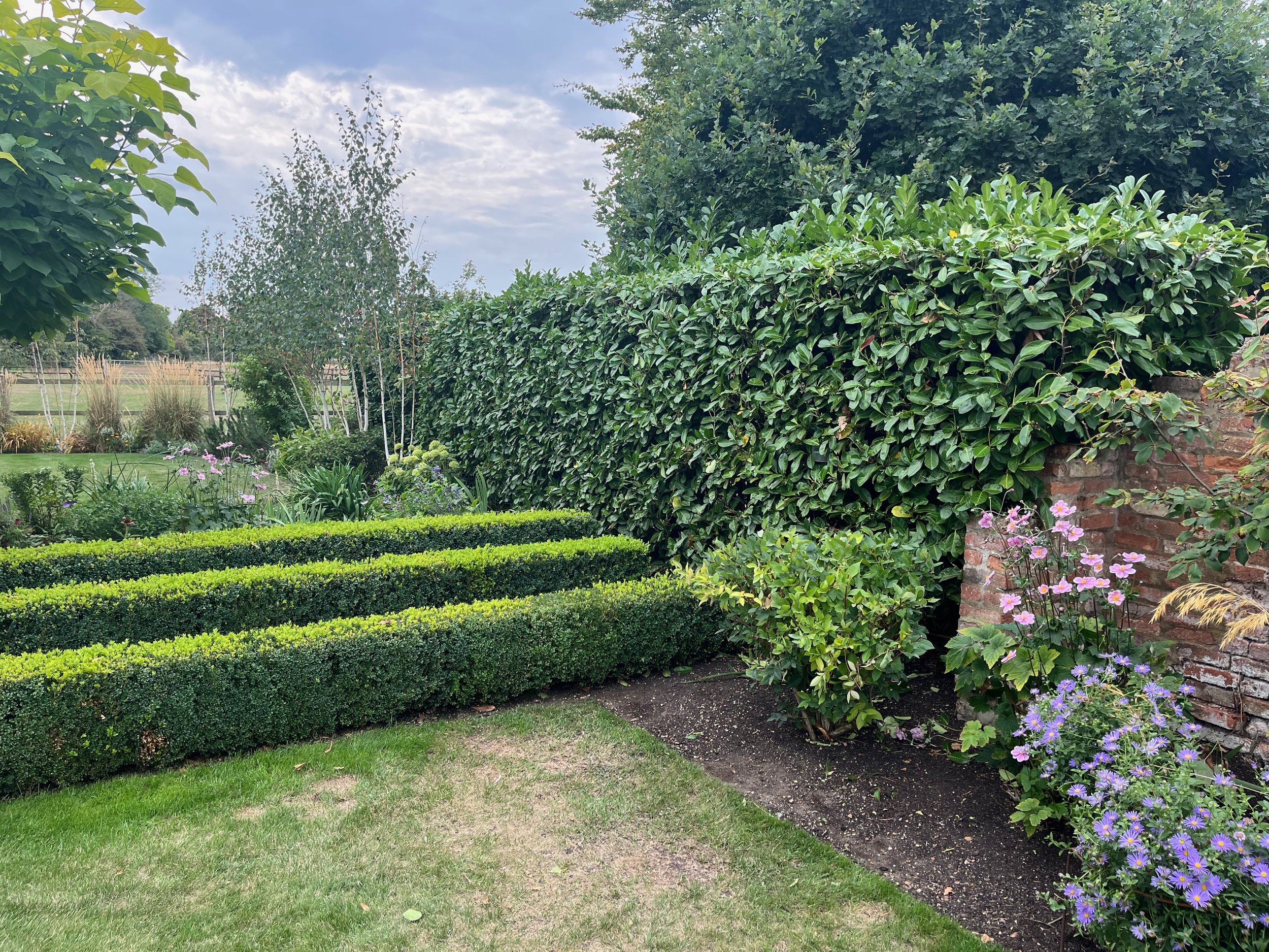 A well-maintained garden with trimmed hedges, flowering plants, and trees under a partly cloudy sky.