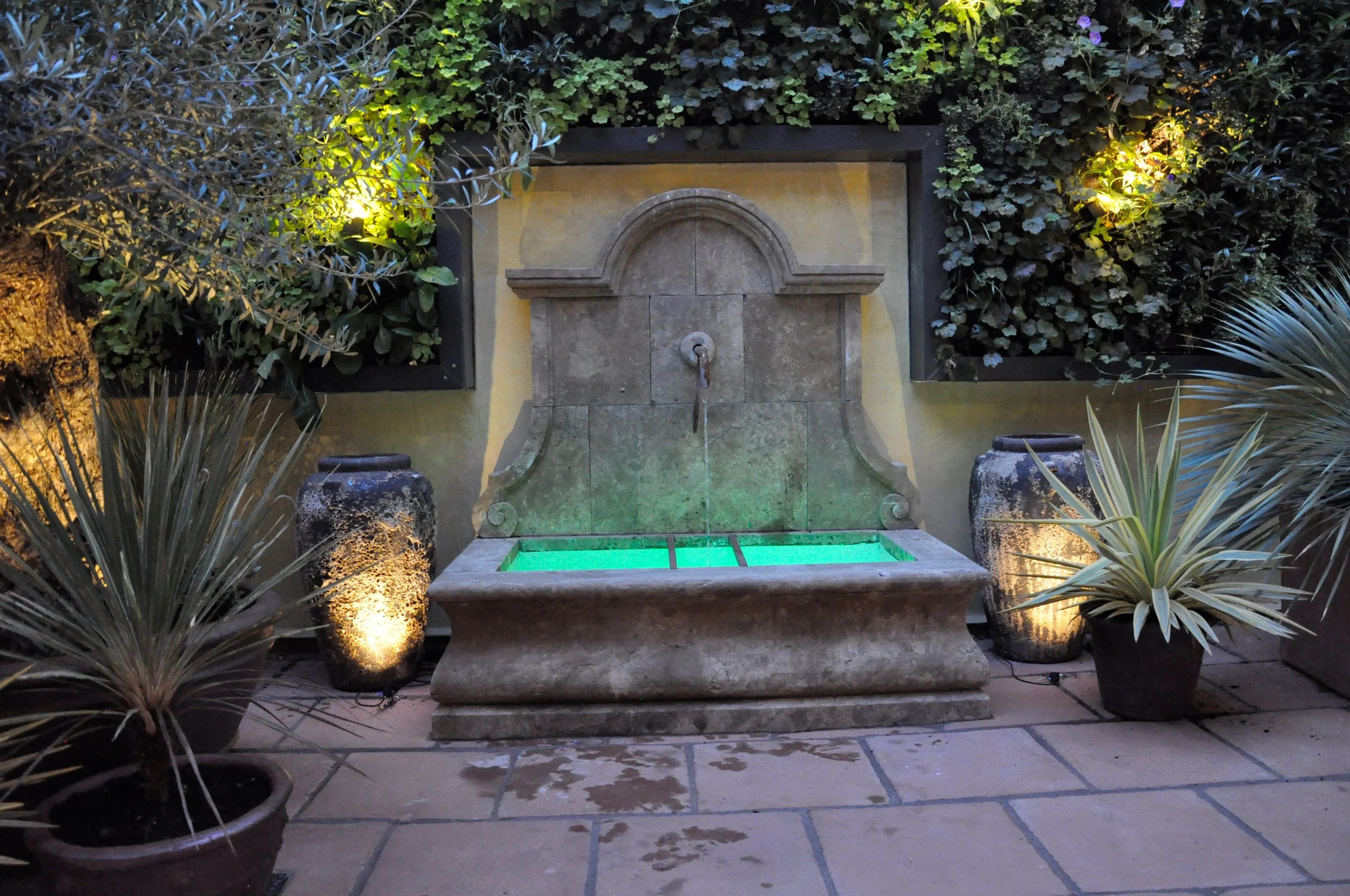 Courtyard garden, outdoor fountain with water illuminated green at night, surrounded by potted plants and greenery, with landscape lighting.