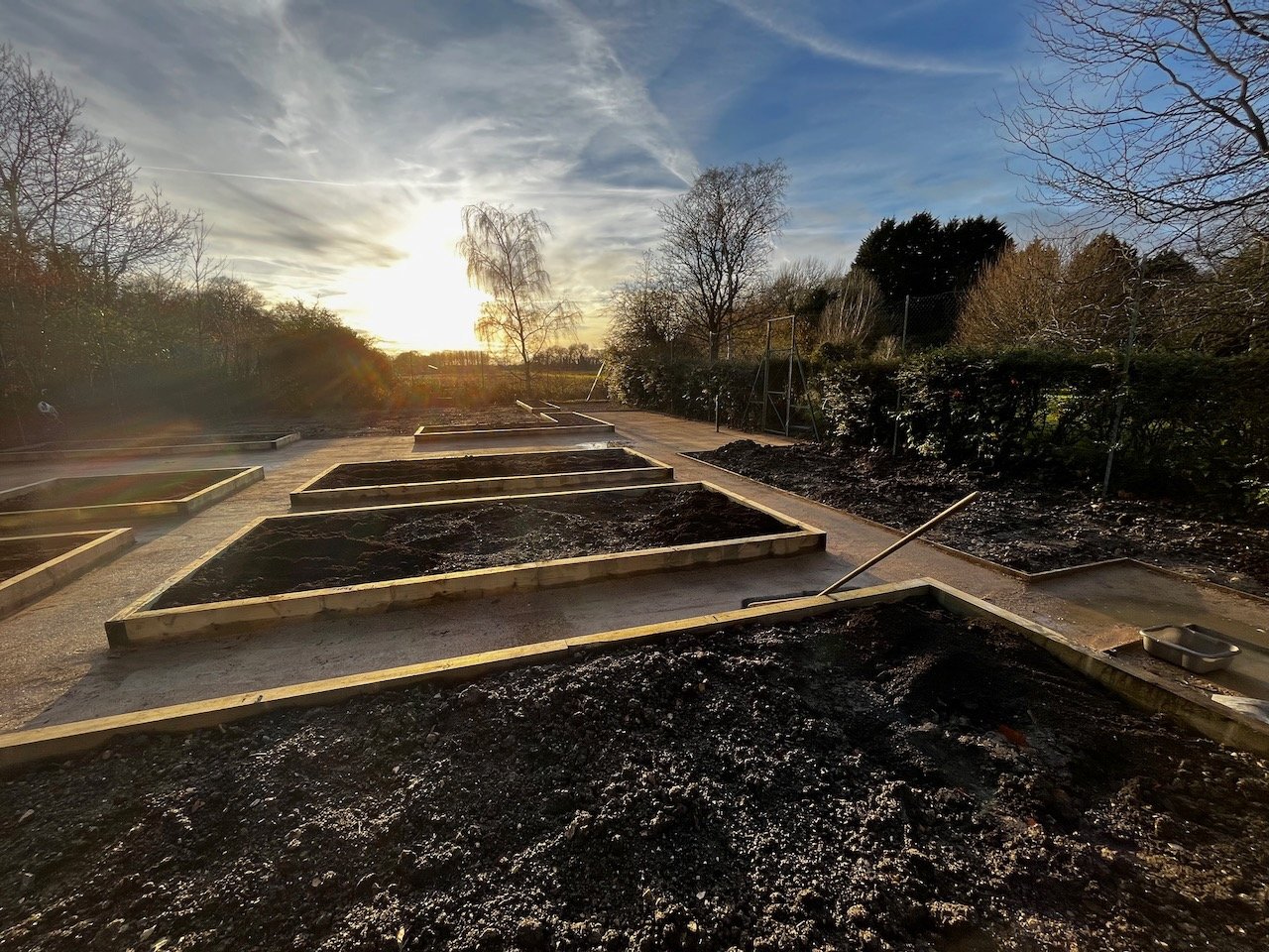 Empty garden bed frames in a garden during late afternoon with the sun setting, trees without leaves, and a cloudy sky.