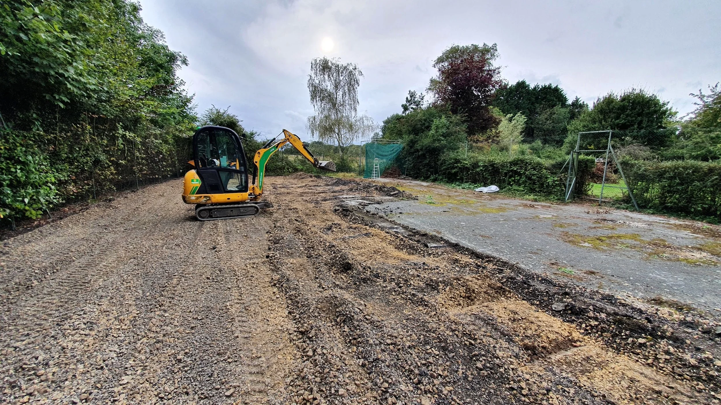 A small yellow and green excavator on a gravelly surface outdoors, with trees and a green netted structure in the background on an overcast day.