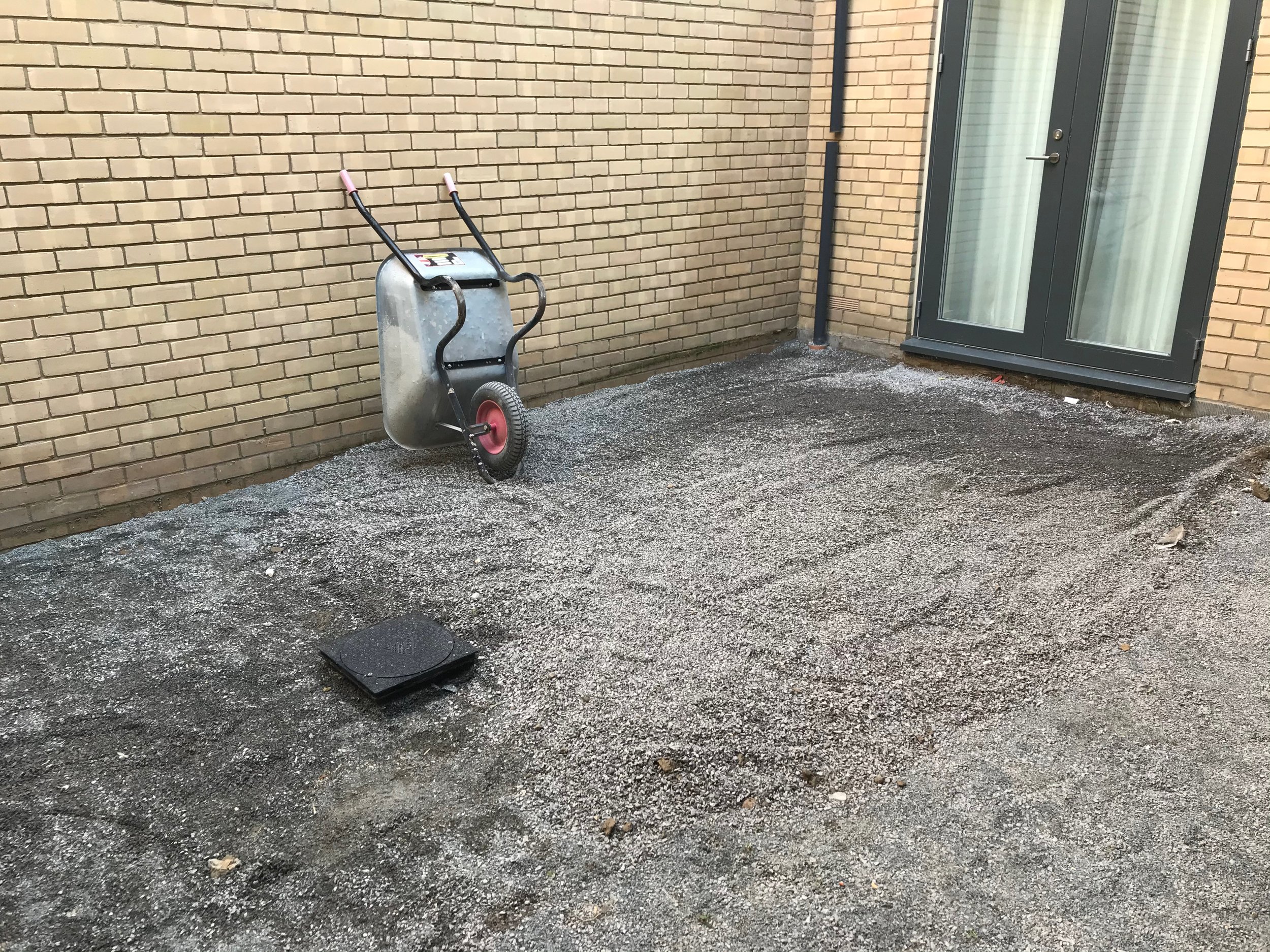 A wheelbarrow with pink handles is on a gravel area outside a brick building near a glass door.
