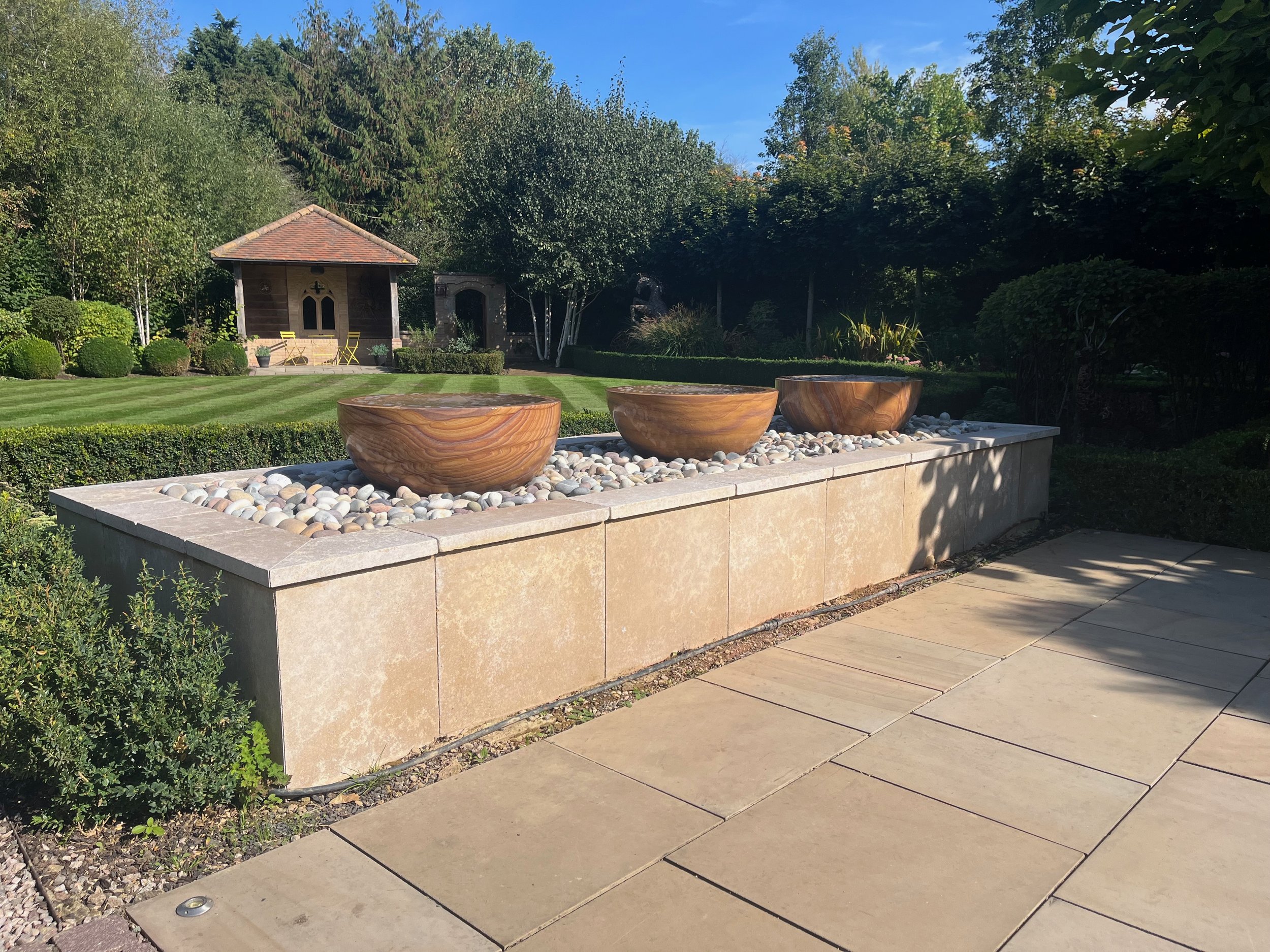 A landscaped backyard with a small wooden shed, outdoor furniture, a raised flower bed with three large wooden bowls, a neatly trimmed hedge, and a lush green tree line under a clear blue sky.