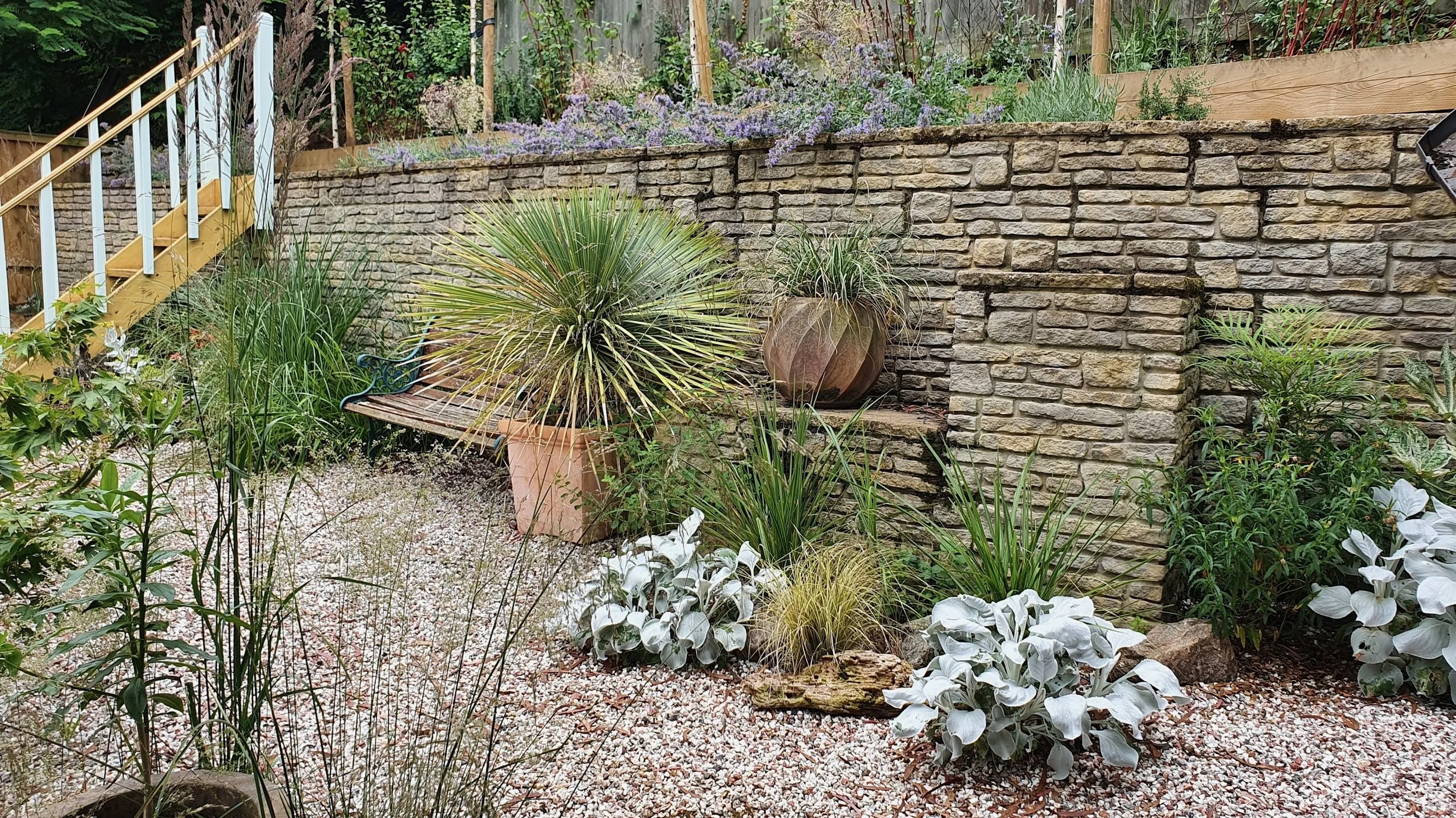 A landscaped garden with a stone retaining wall, various plants, white flowers, potted plants, a wooden bench, and a staircase with a railing on the left side.
