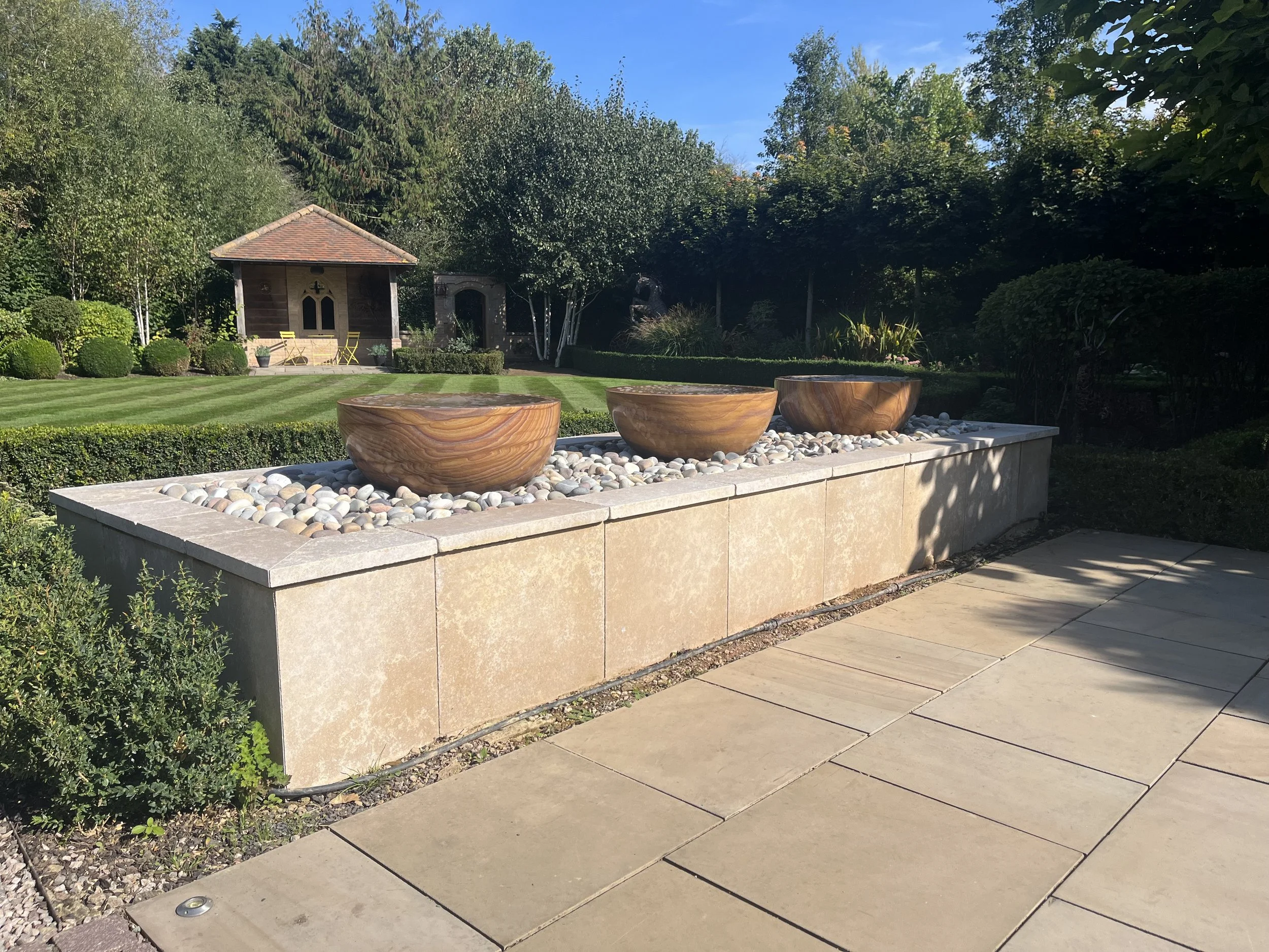 A landscaped garden with a neatly mowed lawn, three large wooden planters, a stone paved border, and a small wooden structure surrounded by lush green trees and shrubs.