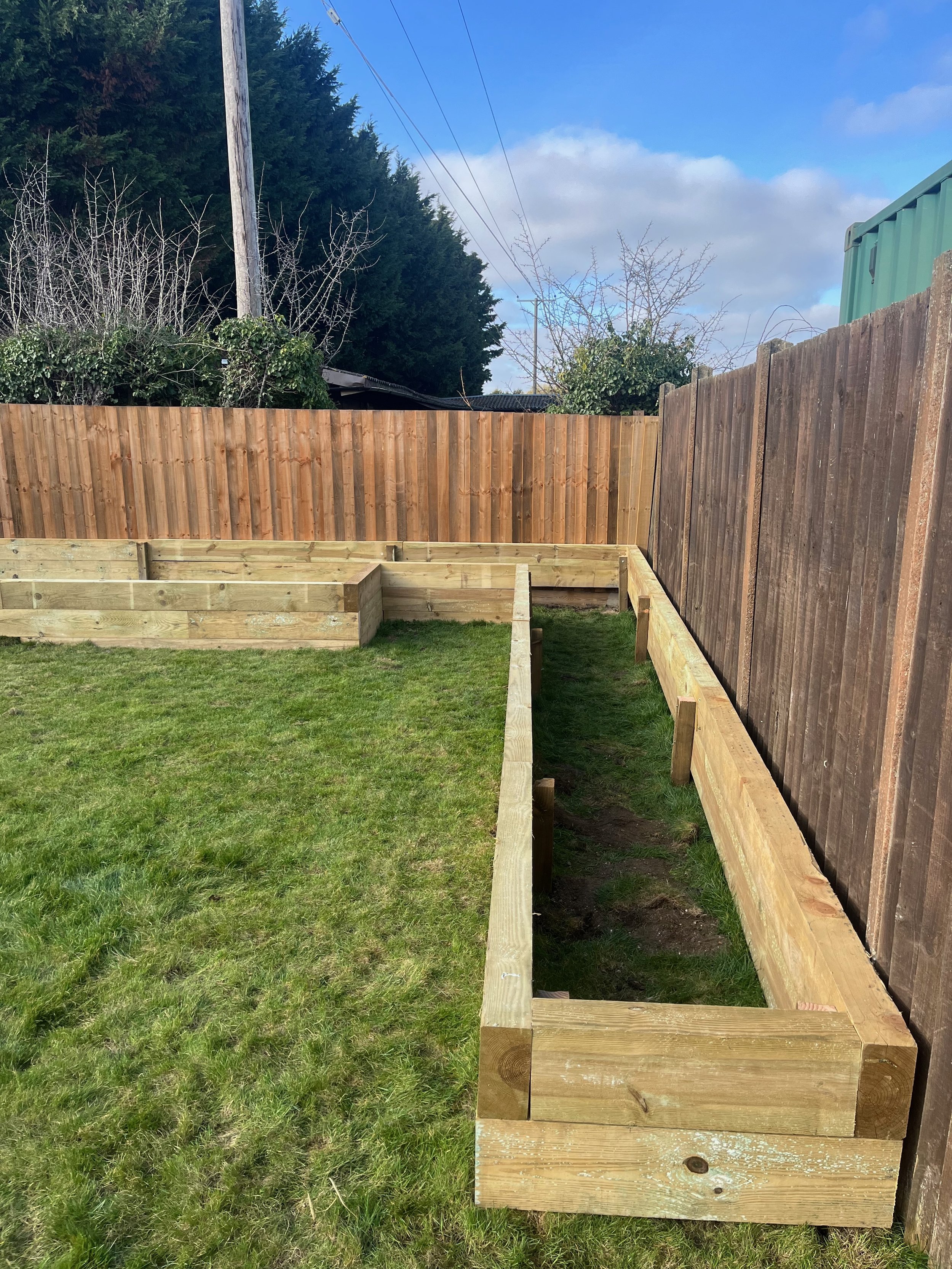 In the backyard, a wooden frame for a garden bed is being built, with one side completed and the other side still under construction, surrounded by green grass. A brown wooden fence and trees are visible in the background, with a utility pole and pow