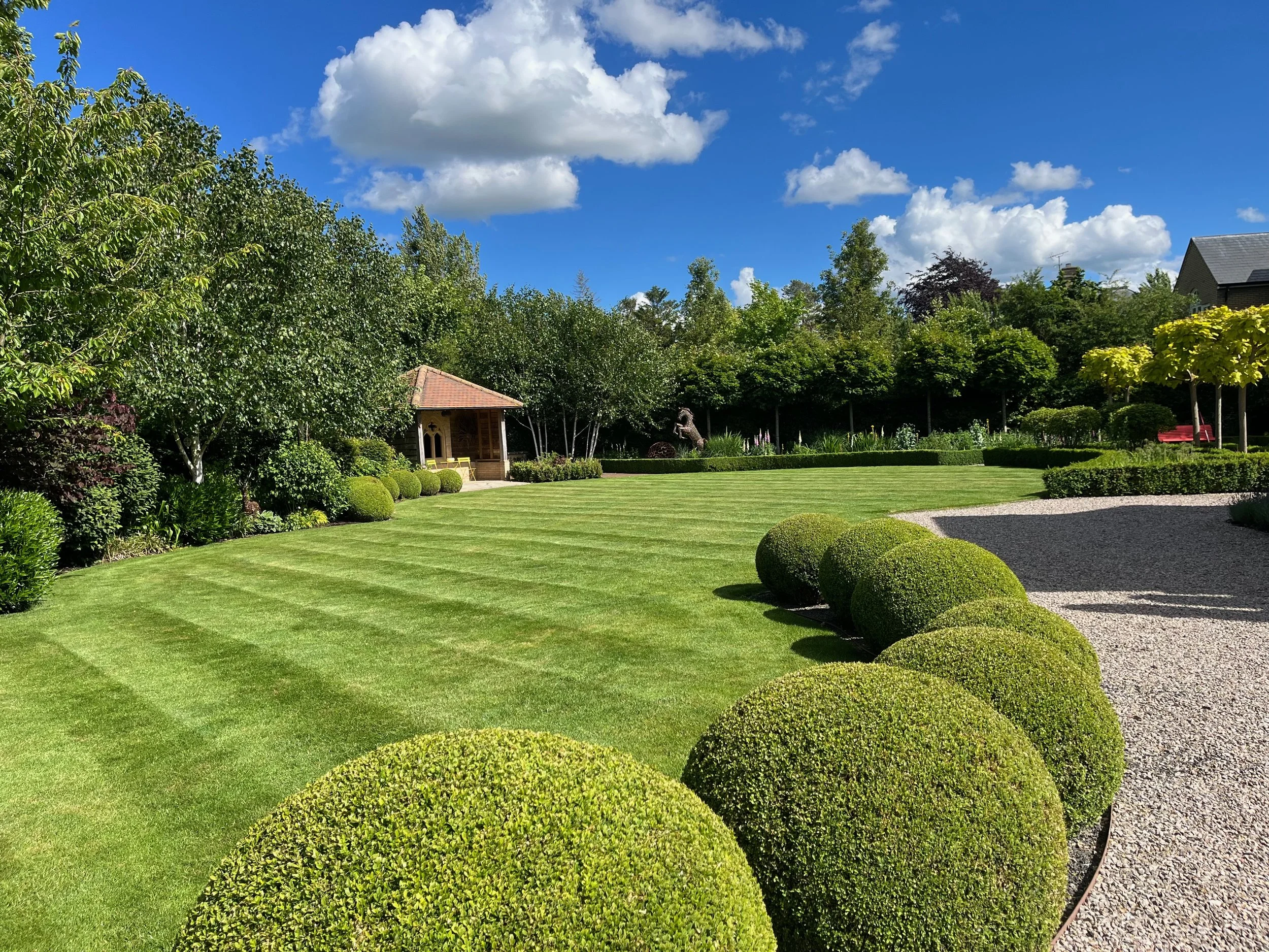 A backyard garden with a well-manicured lawn, bushes, trees, a small shelter, and a house, under a blue sky with fluffy white clouds.