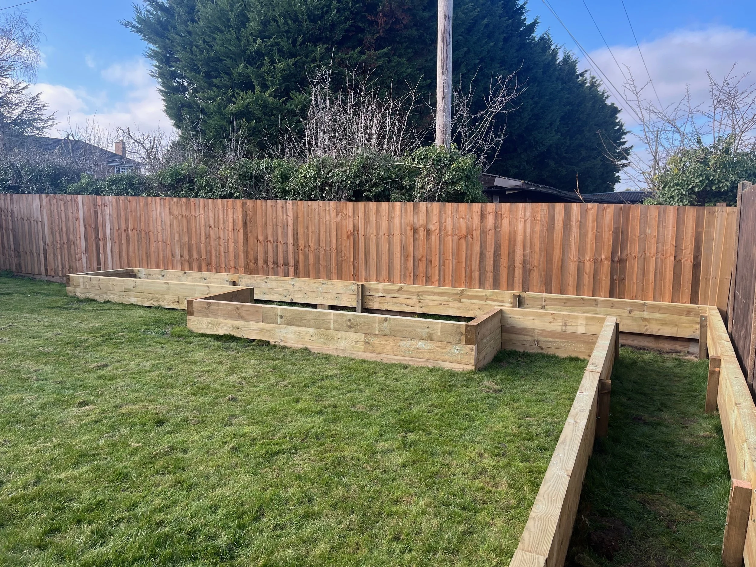 A backyard with a grassy lawn, a wooden fence, and a newly built wooden garden bed framework. Behind the fence are trees and a house partially visible.