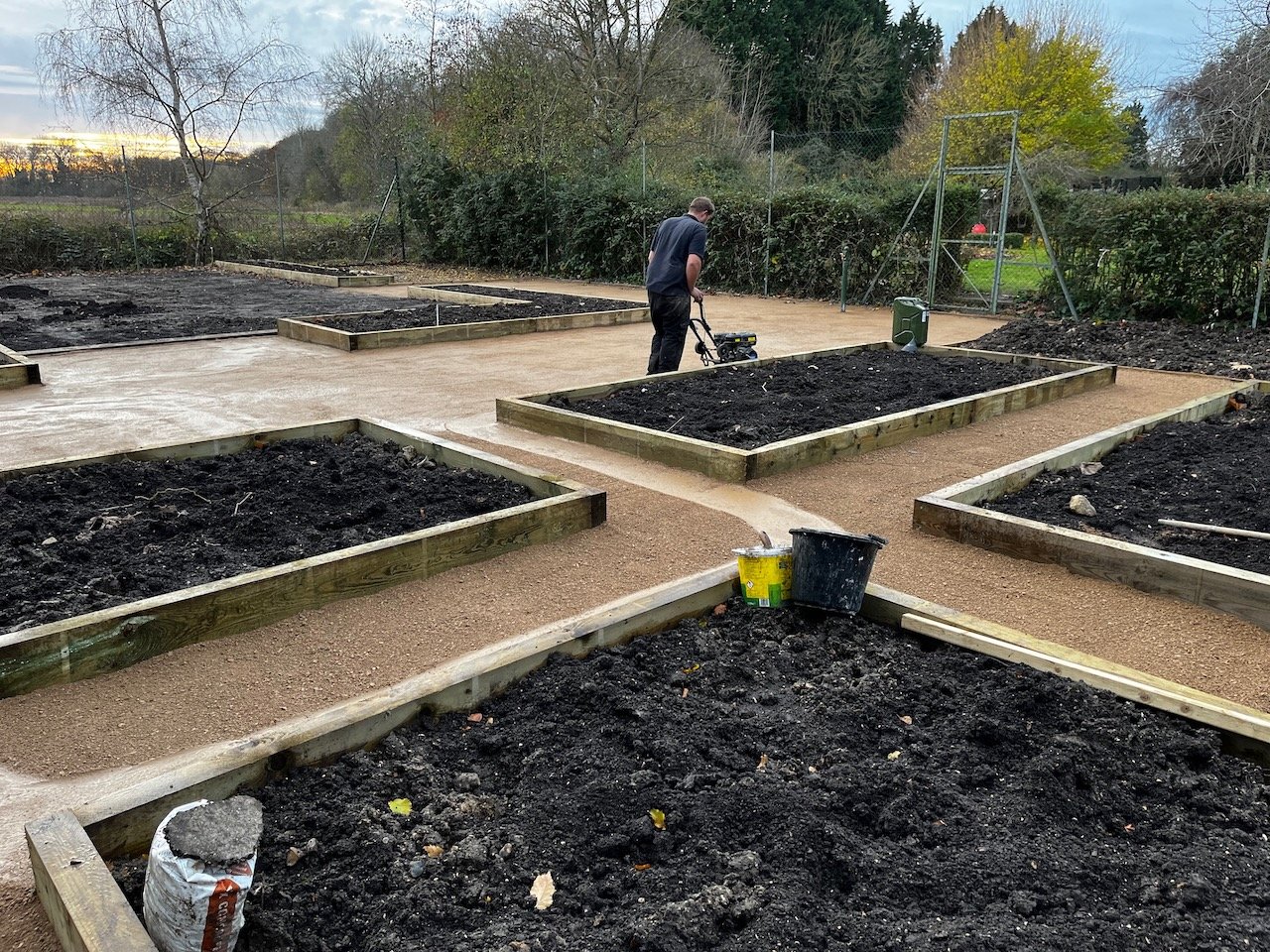 A person is working on a community garden or allotment with raised beds, tiling the pathways between the beds, in an outdoor setting with trees and a fence in the background.