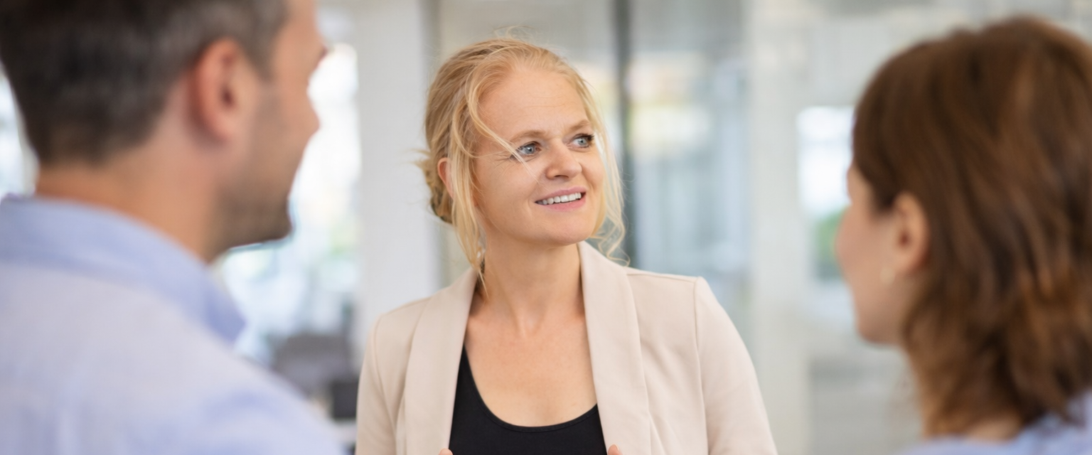 Drei Menschen im Gespräch in einem modernen Büro, eine Frau schaut freundlich und aufmerksam, die anderen beiden sind teilweise sichtbar.