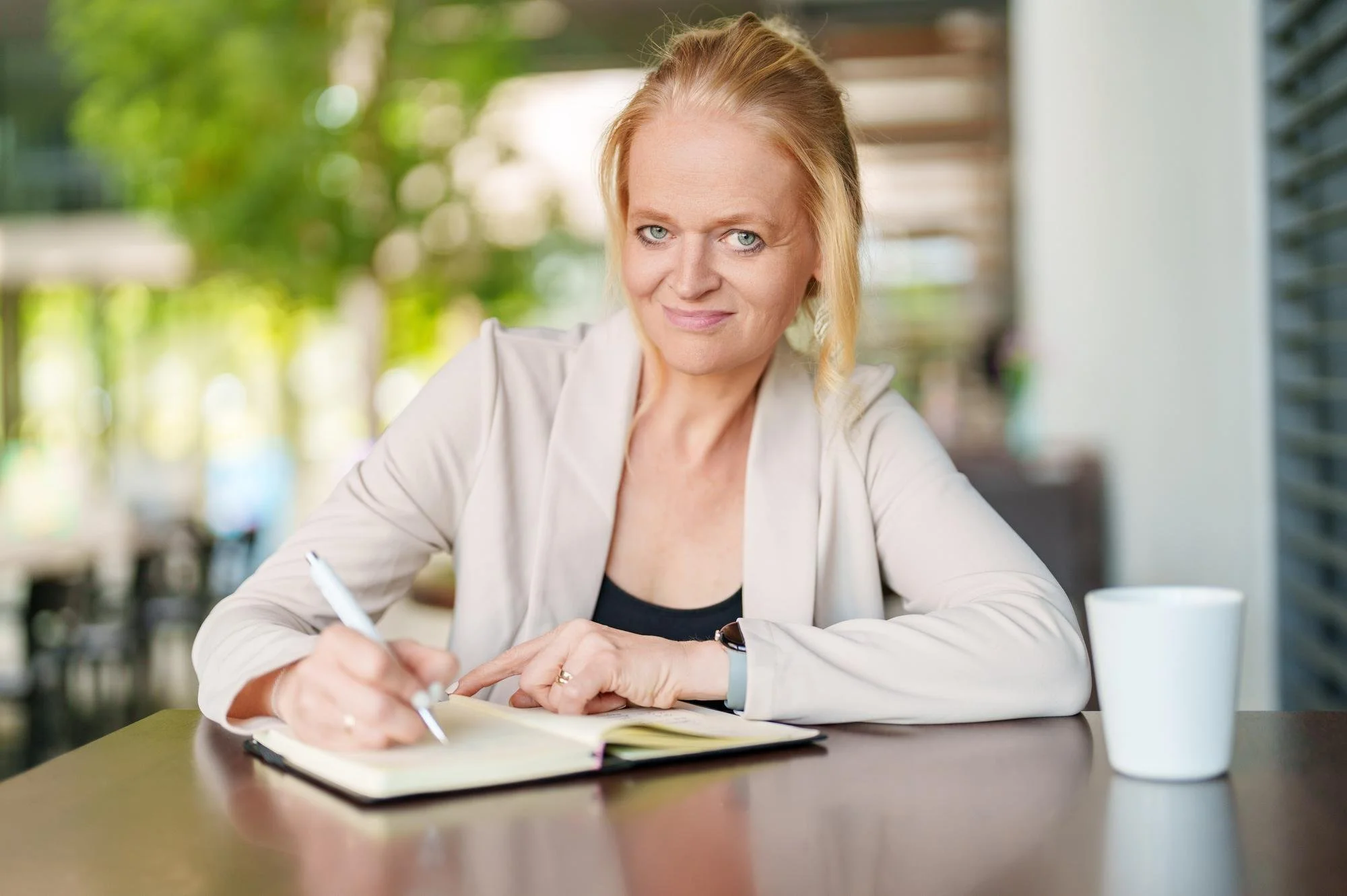 Eine blonde Frau sitzt in einem hellen Raum mit großen Fenstern, schreibt in ein Notizbuch und schaut lächelnd. Es ist eine angenehme, natürliche Lichtstimmung. Neben ihr steht eine weiße Kaffeetasse.