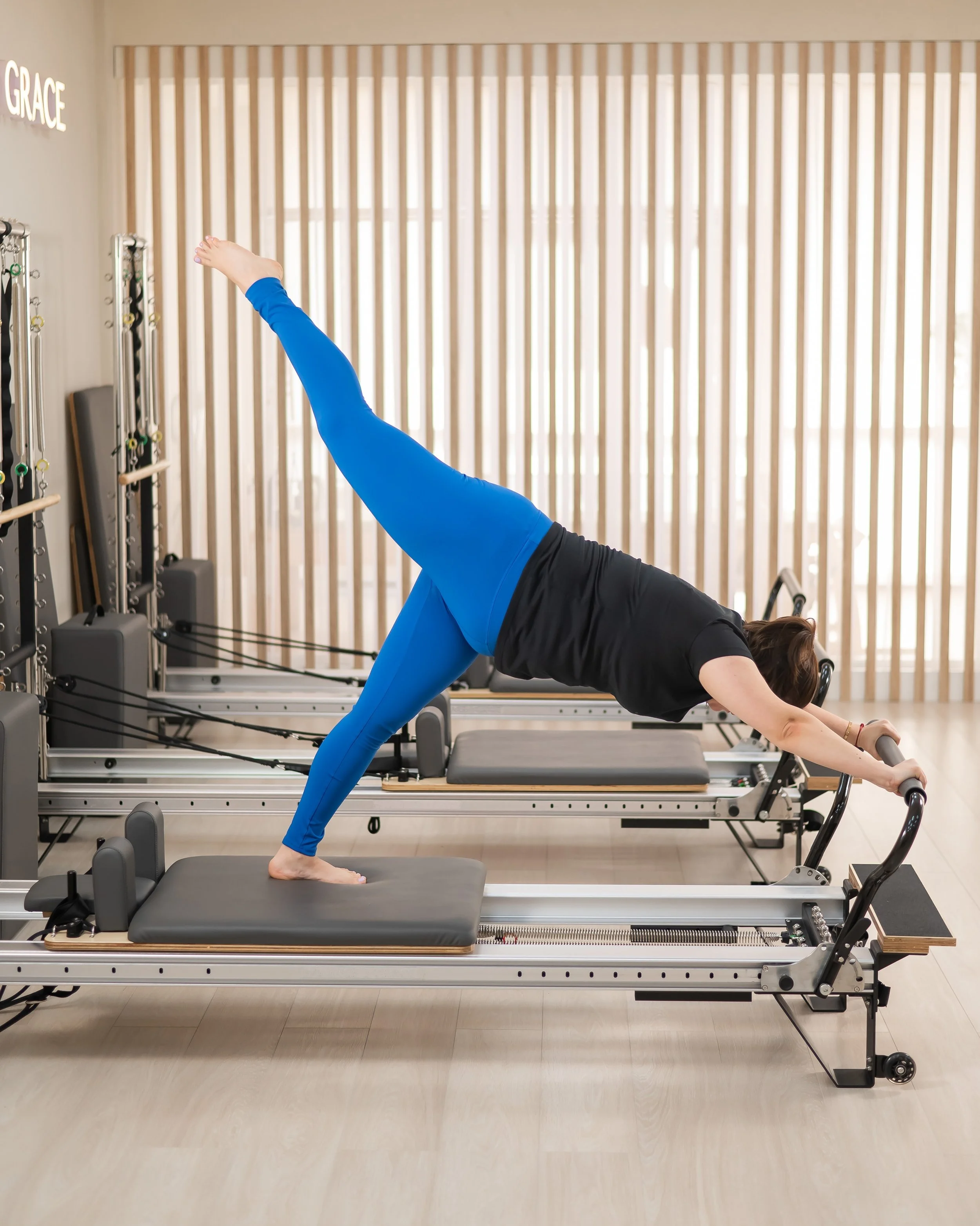 A woman in a blue tights performing a Pilates exercise on a reformer machine in a Pilates studio, with wooden blinds.