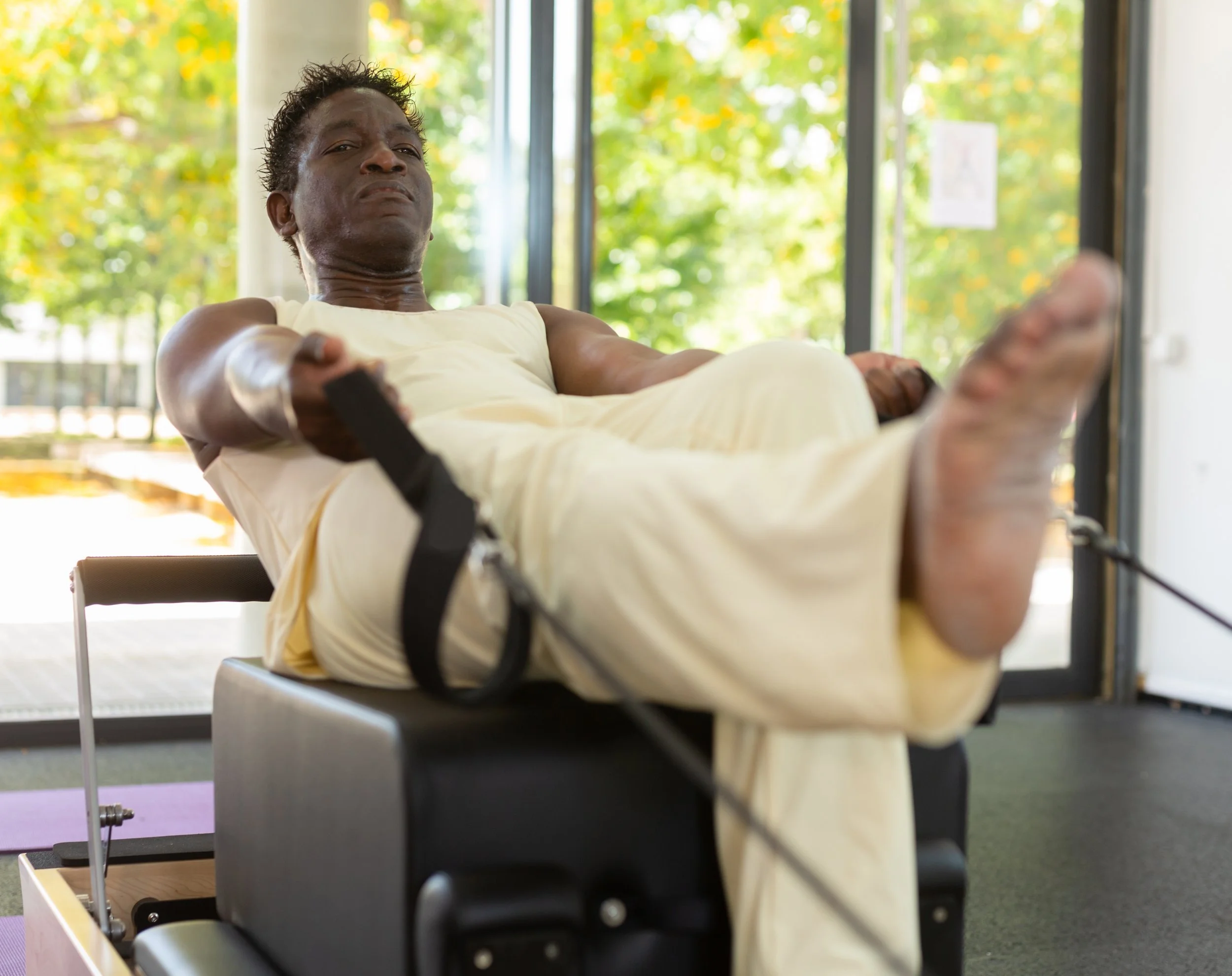 A man doing a roll down on a Pilates reformer with one leg up, in a room with large windows showing trees outside.