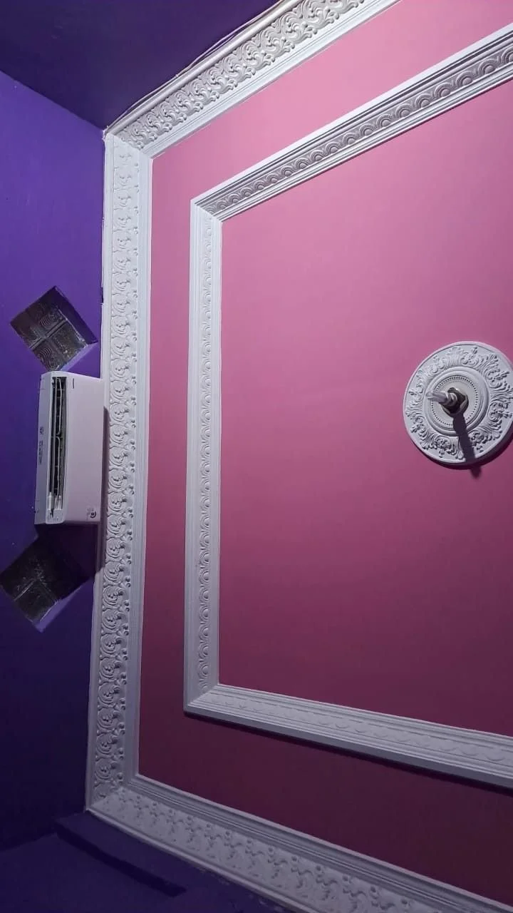 Une pièce avec mur rose et une partie de mur violet, décorée avec des moulures blanches ornées, et une unité de climatisation fixée au mur violet avec des surfaces métalliques réfléchissantes. Une prise électrique est visible sur le mur rose.
