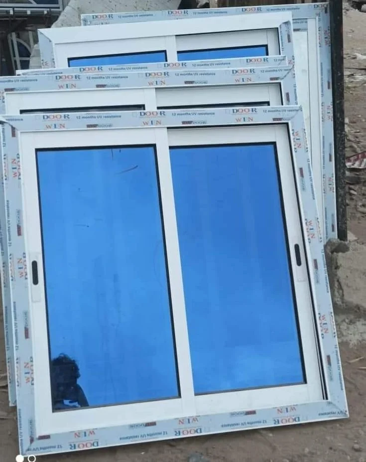 Fenêtre à cadre blanc avec deux volets en verre, posée sur le sol avec un cadre supplémentaire en arrière-plan. Encadrement en plastique avec étiquettes et inscriptions en anglais.