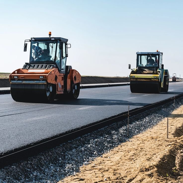 Deux rouleaux compresseurs asphaltent une route sur un terrain plat, avec un ciel clair.