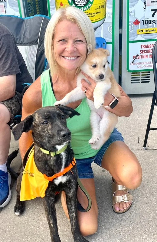 A woman smiling and kneeling while holding a small light-colored puppy in her right arm, and a black dog with a yellow vest sitting beside her on the ground. They are outdoors, near what appears to be a recycling or waste disposal area with signs in the background.