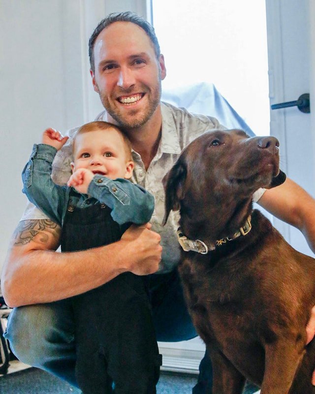 A man, a baby, and a dog posing together indoors. The man has a beard and is smiling, holding the baby, who is smiling and raising a hand. The dog is a large brown Labrador Retriever, sitting with a calm expression.