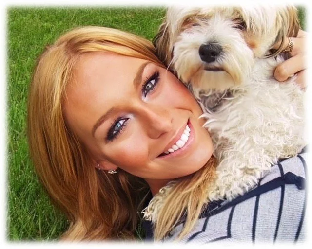 A smiling woman with blonde hair lying on green grass holding a small, fluffy white dog close to her face.