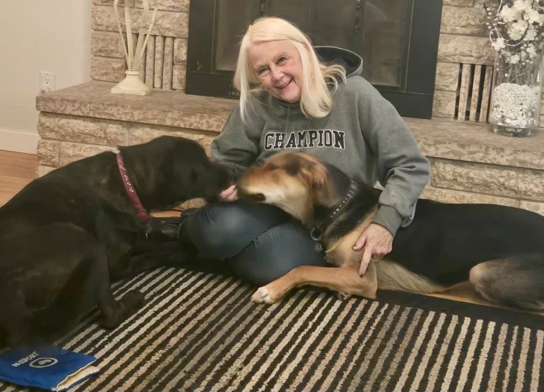 An elderly woman with blonde hair sitting on a striped rug in front of a brick fireplace, smiling, with two dogs. One dog is black and tan, lying on the woman’s lap; the other is black and brown, lying next to her, with her arm around it.