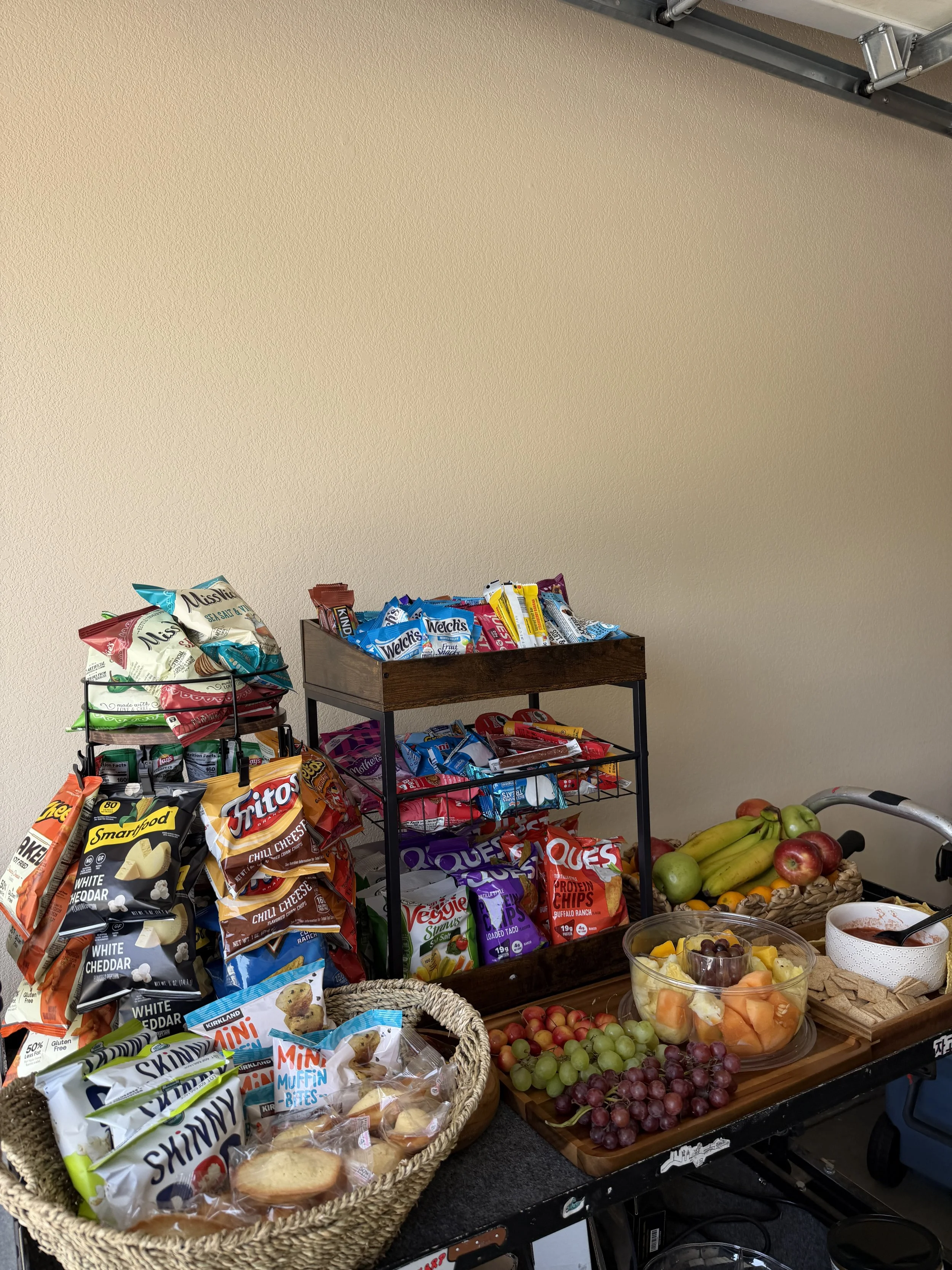 A table and shelves filled with snack bags, fruits, and other food items in a kitchen or pantry area.