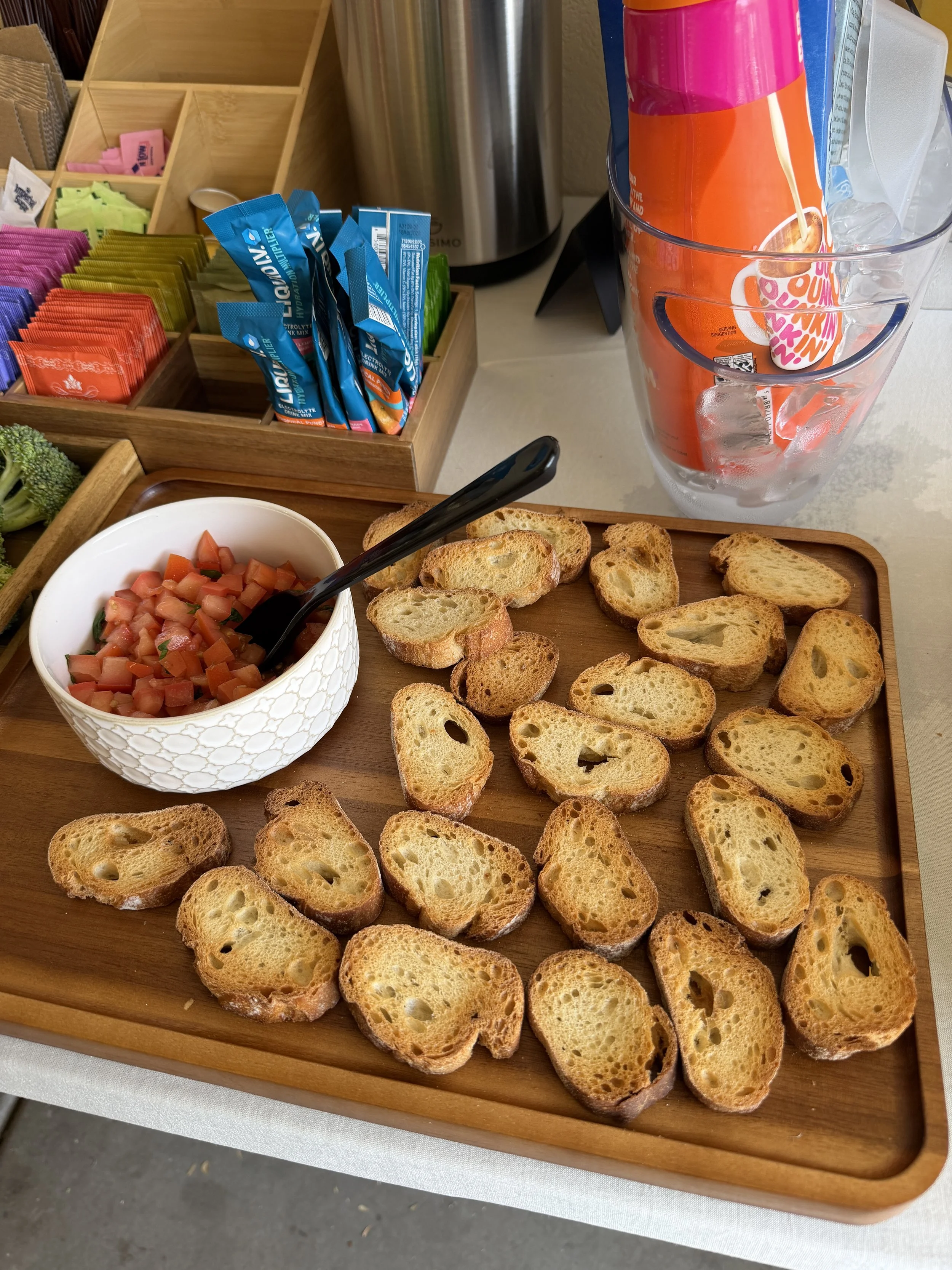 A wooden tray with sliced toasted baguette pieces, a bowl of chopped tomatoes with a black serving spoon, a glass with ice and a pink bottle inside, packets of sweetener and sugar in a wooden organizer, a large metal container, and a disposable spoon, in a breakfast or buffet setup.