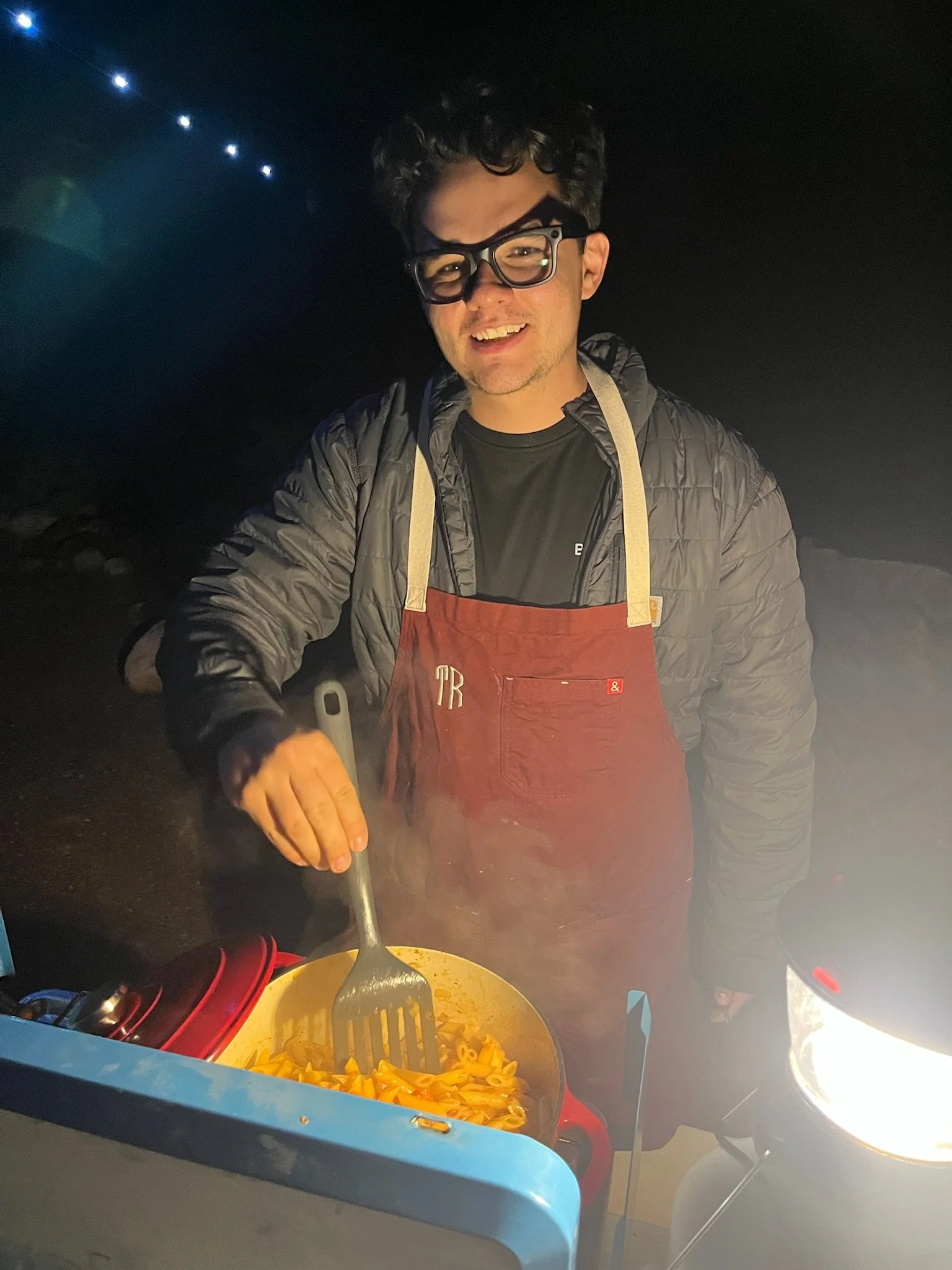 Person cooking pasta in a large blue pot at night, wearing glasses, a black shirt, and a gray jacket, with a grill and some lights visible in the background.