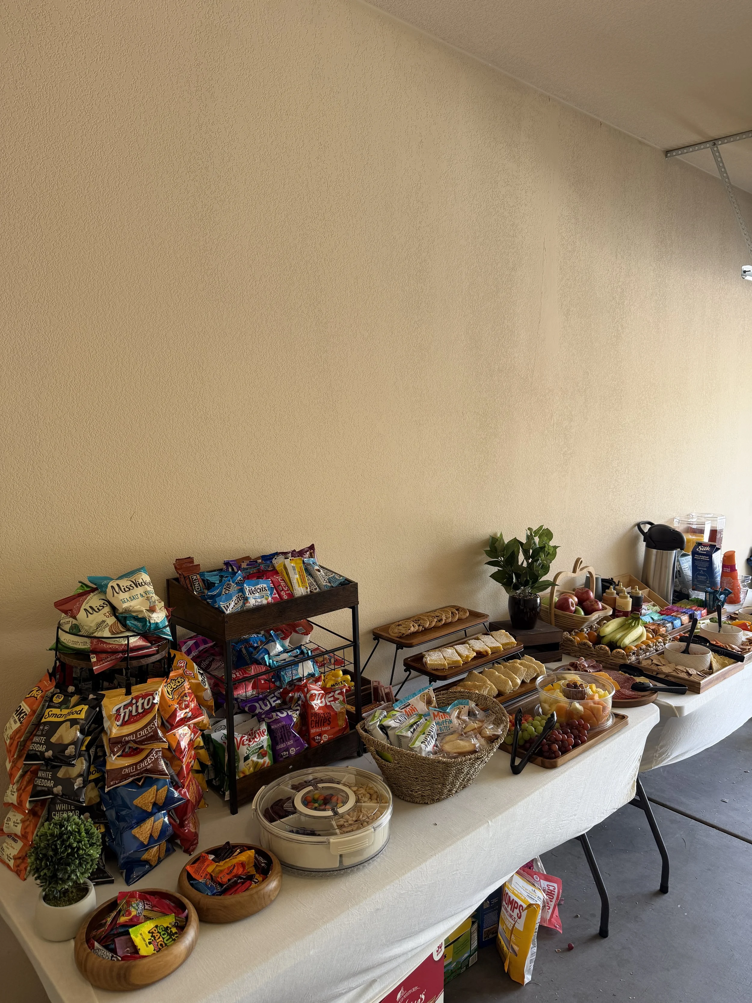 A table set up with snacks and drinks for a gathering, featuring chips, cookies, fruit, and condiments, with a wall in the background.