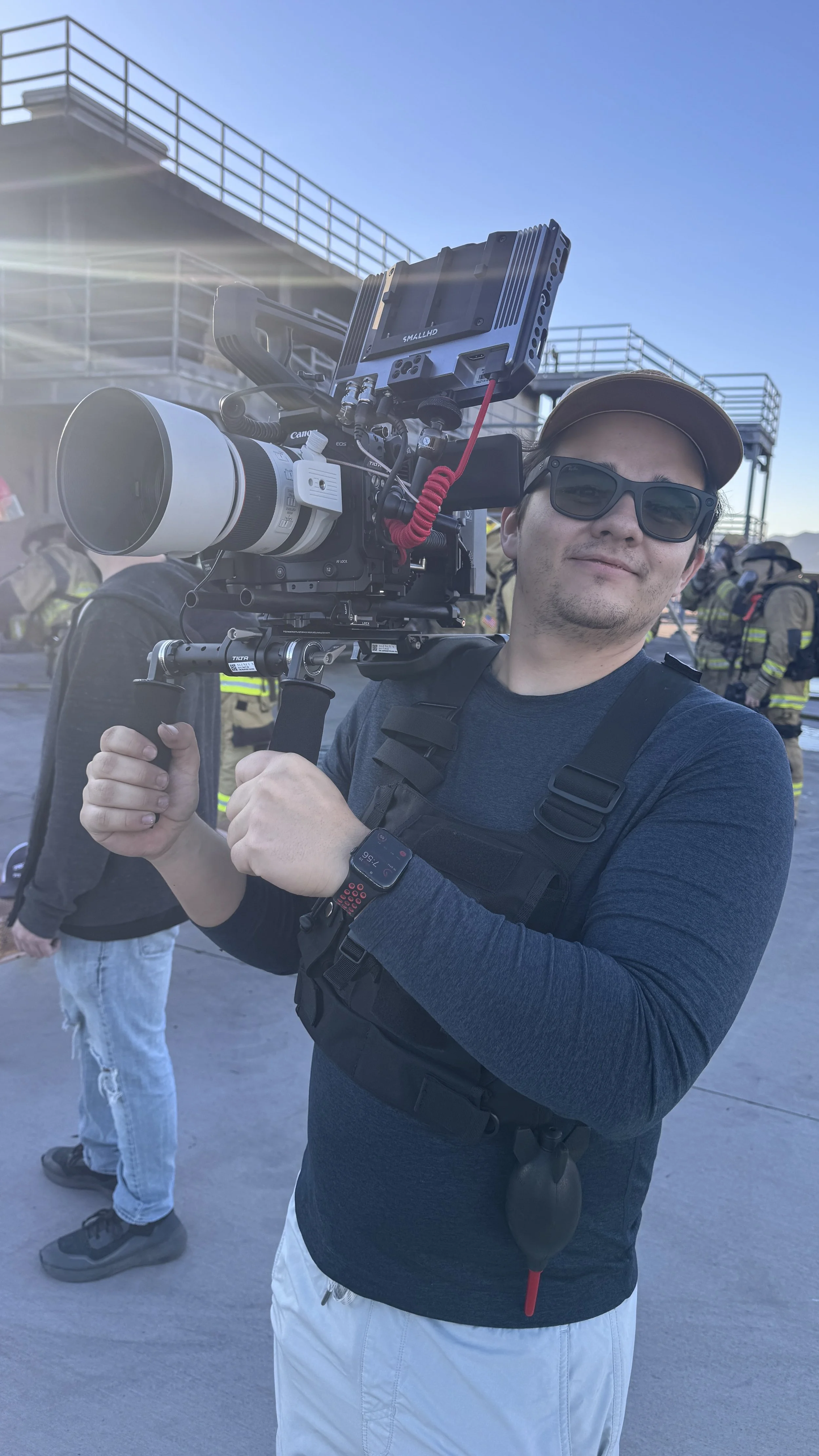 A man wearing sunglasses and a cap is holding a professional video camera with a large lens and external monitor, standing outdoors in bright sunlight with a clear blue sky.