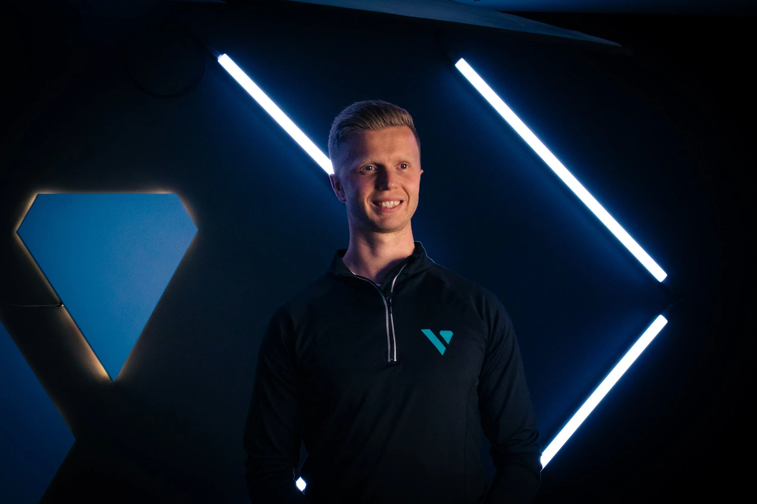 A man with blonde hair wearing a black jacket with a blue logo, standing in front of a dark background with neon blue and white lights arranged in geometric patterns.