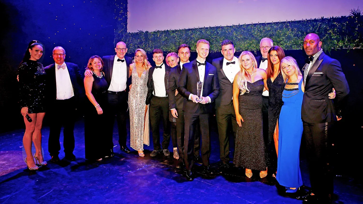 Group of people dressed in formal attire at an awards ceremony, standing on a stage with a dark background and green foliage, some smiling and one person holding a trophy.