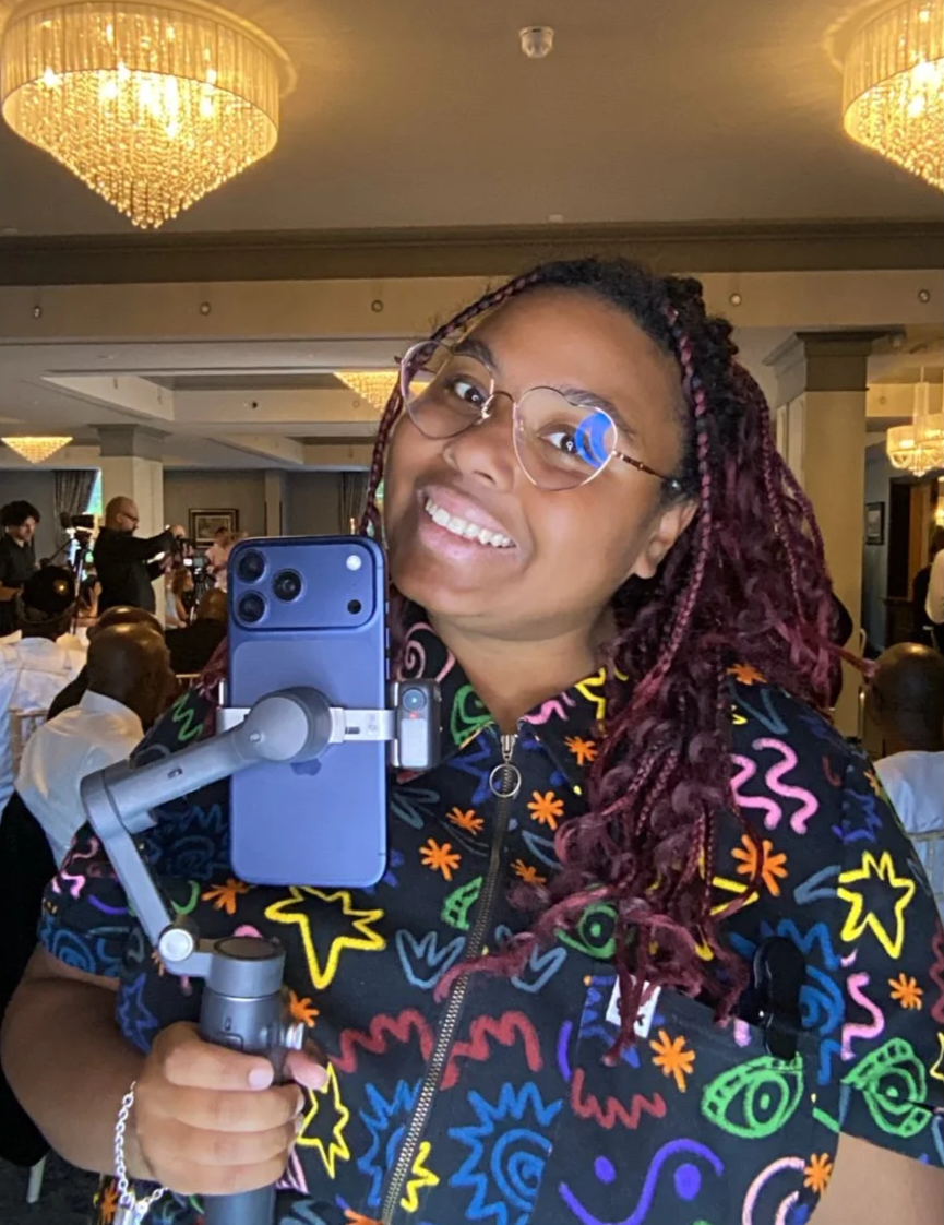 A woman with glasses taking a selfie at a formal event with chandeliers hanging from the ceiling and people in the background.