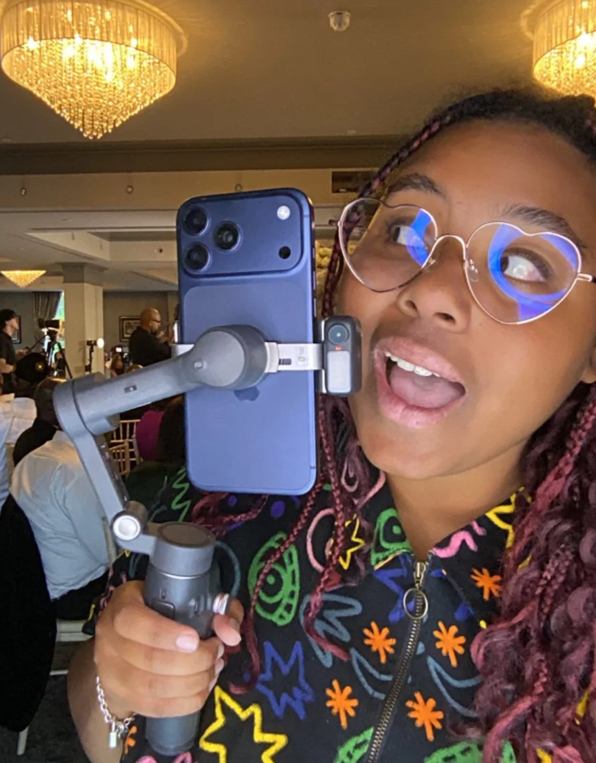 A woman with glasses and braided hair taking a selfie with a smartphone on a stabilizer in a well-lit event space with chandeliers.