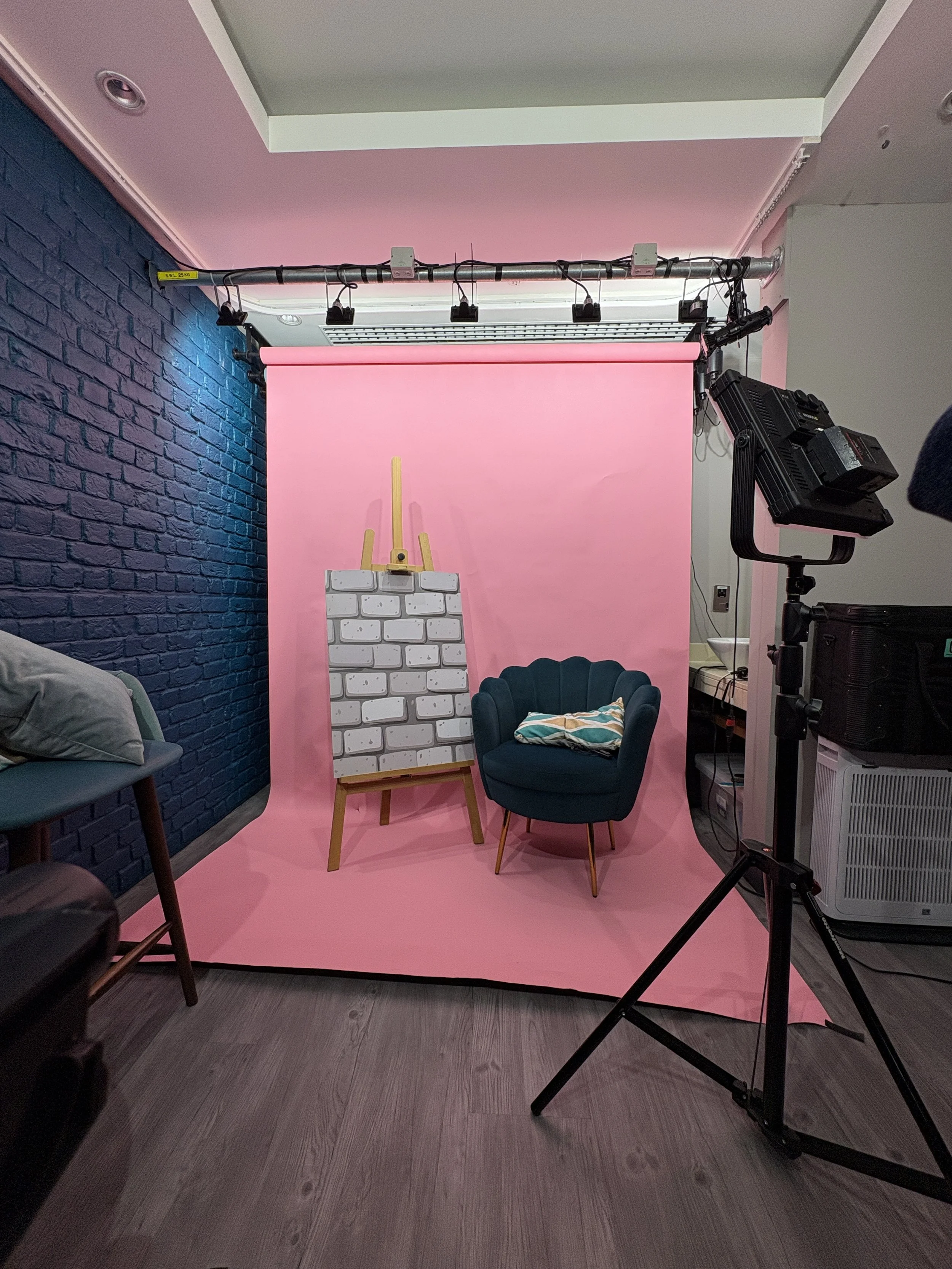 Photo of a photography studio setup with a pink backdrop, an easel with a white brick pattern, a navy blue armchair, and a lighting setup, with a textured blue wall on the left and a gray floor.