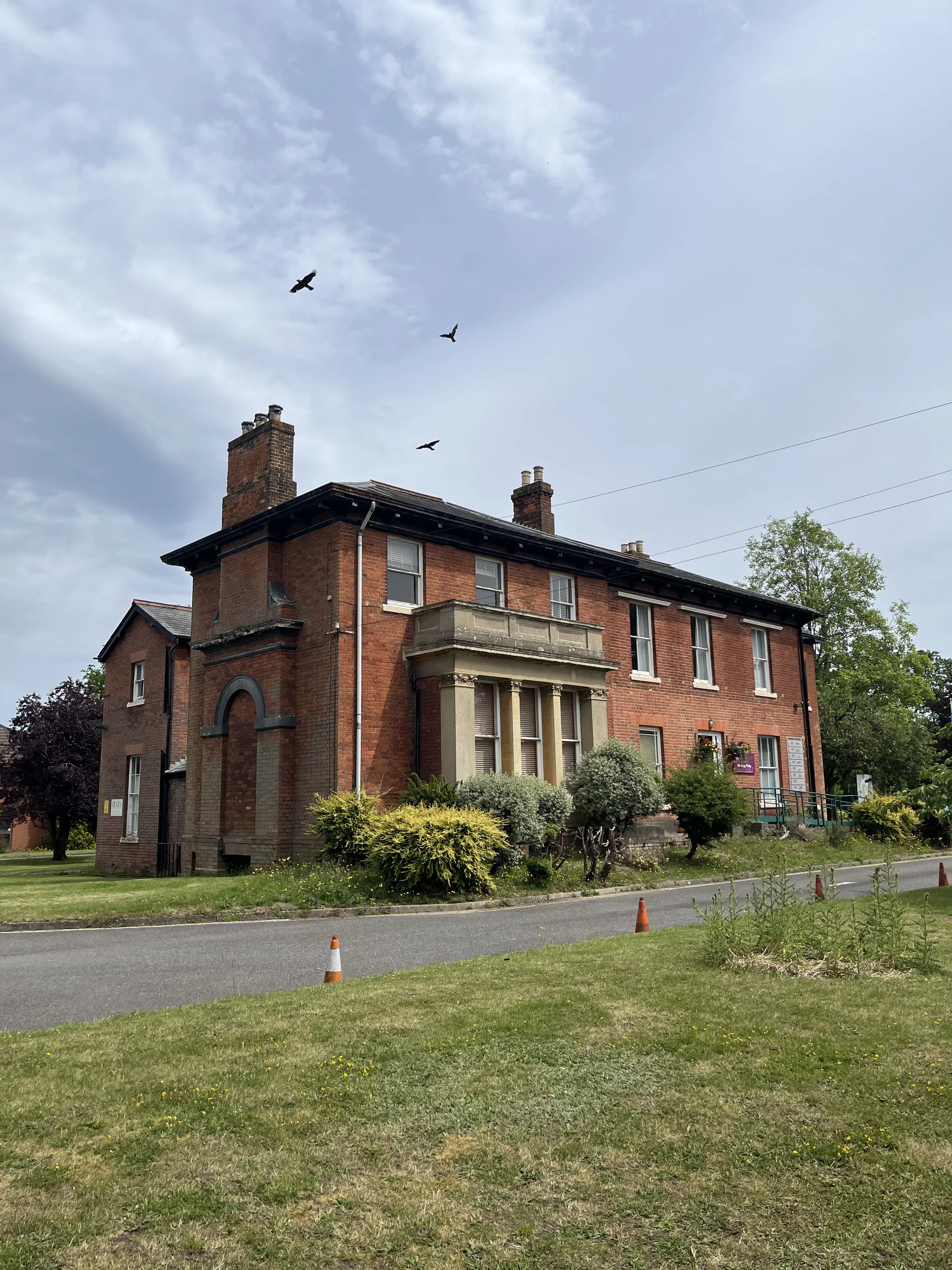 A large brick building with multiple windows and two prominent chimneys, surrounded by a grassy lawn with scattered bushes and trees. The sky above is partly cloudy with three birds flying.