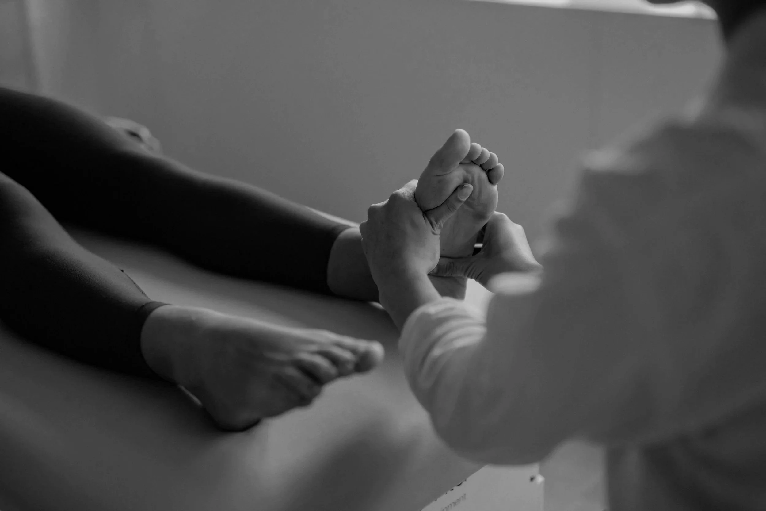 Black and white photo of a person's arm extended, with a healthcare professional holding their hand. The scene appears to be in a medical or clinical setting.
