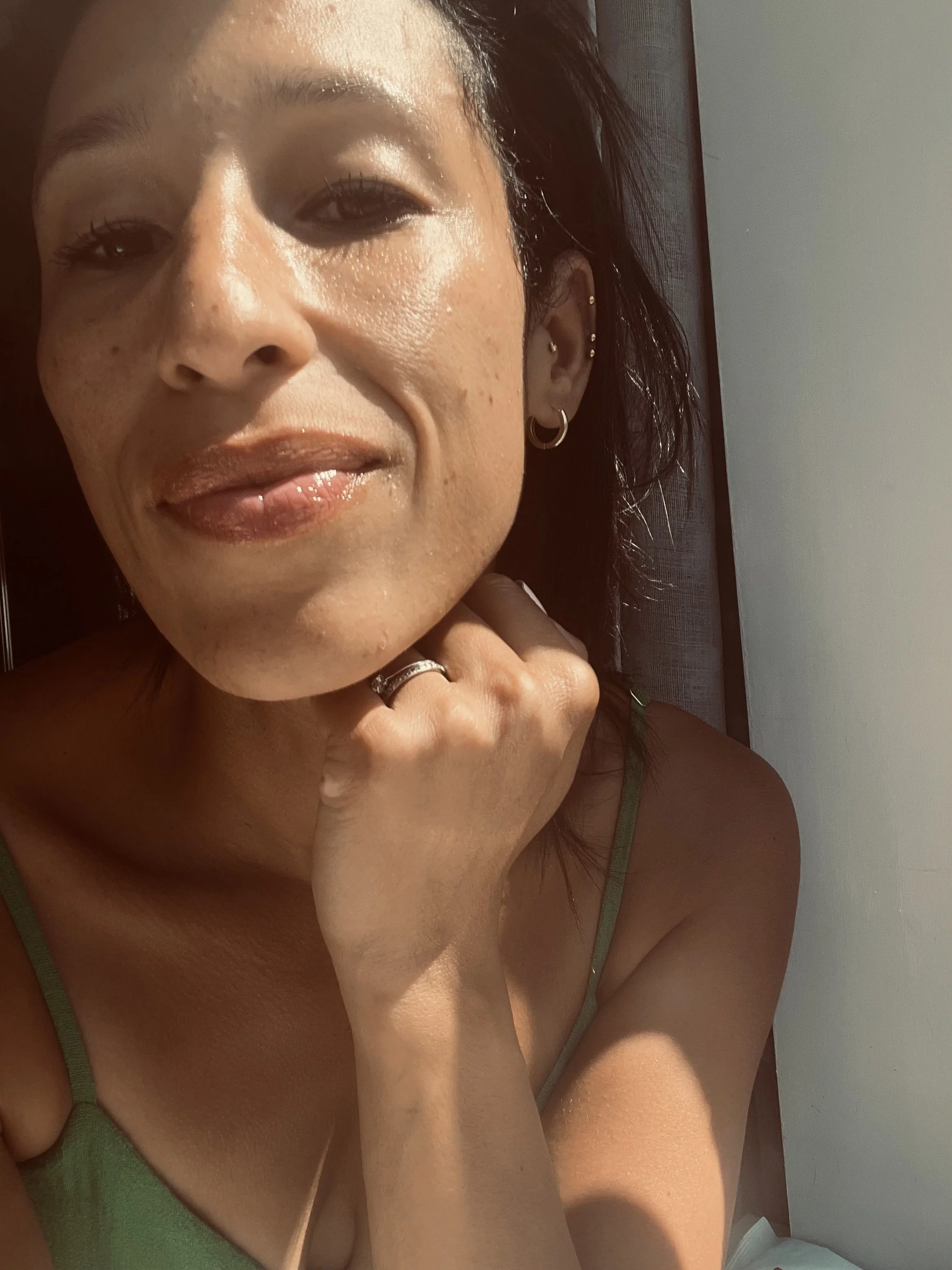 Close-up of a woman with dark hair, wearing makeup, earrings, and a green strap top, resting her chin on her hand in front of a white and brown background.